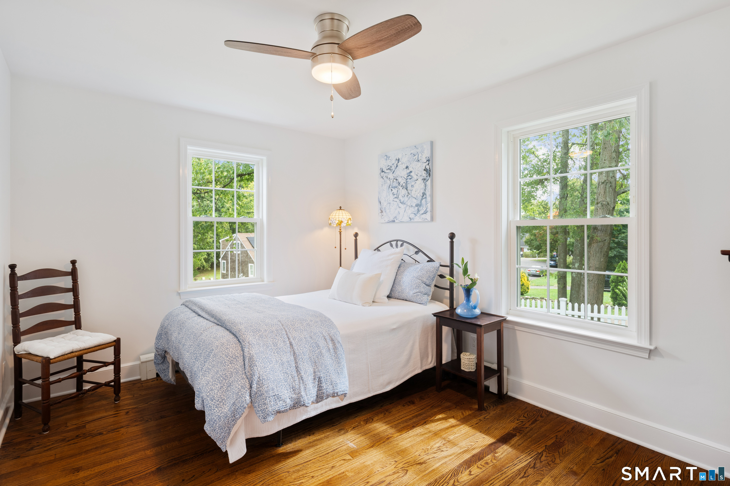 Bright bedroom with pale blue bedding, dark wood floors, and two large windows overlooking greenery.