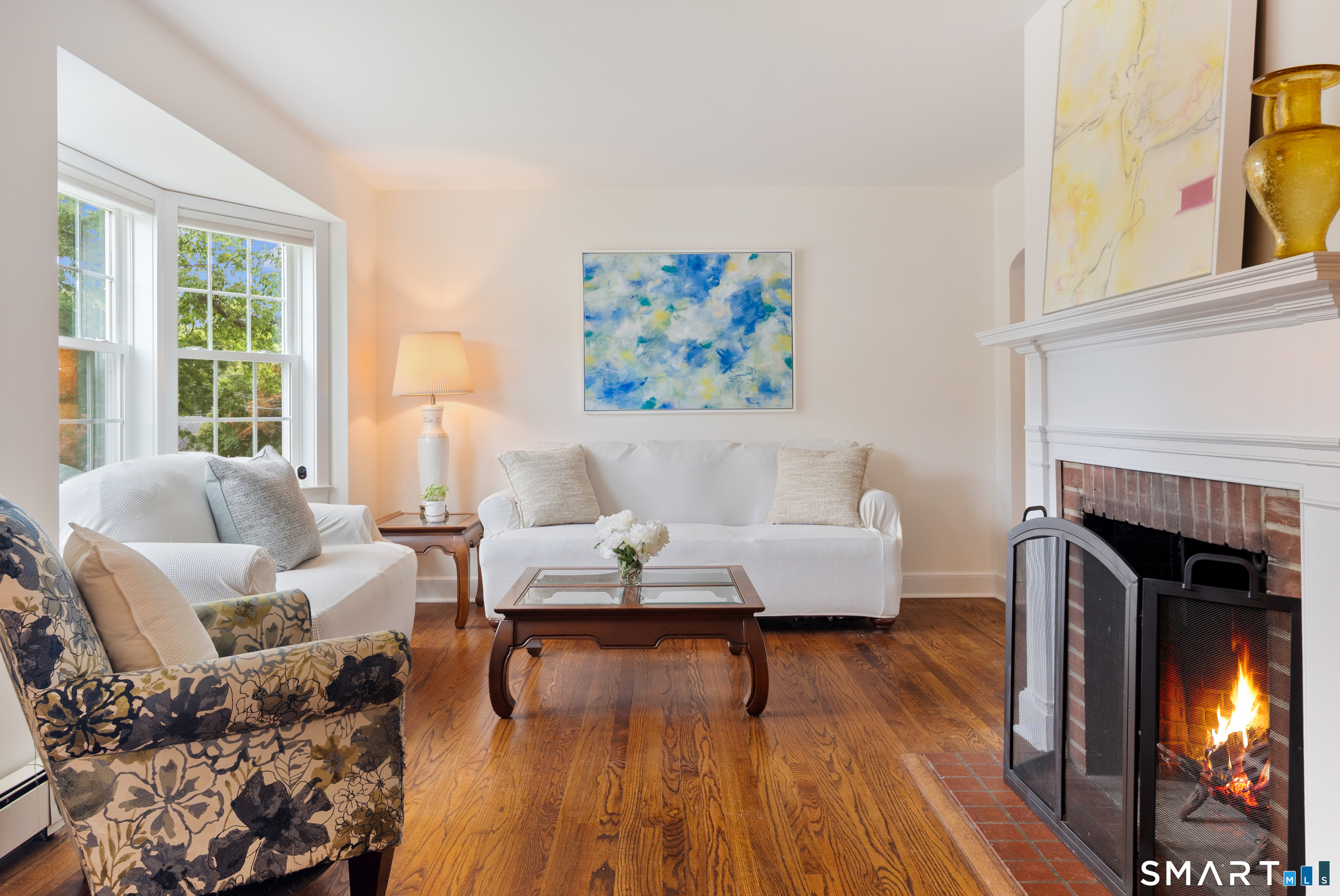 Living room with hardwood floors, large windows, and a lit brick fireplace.