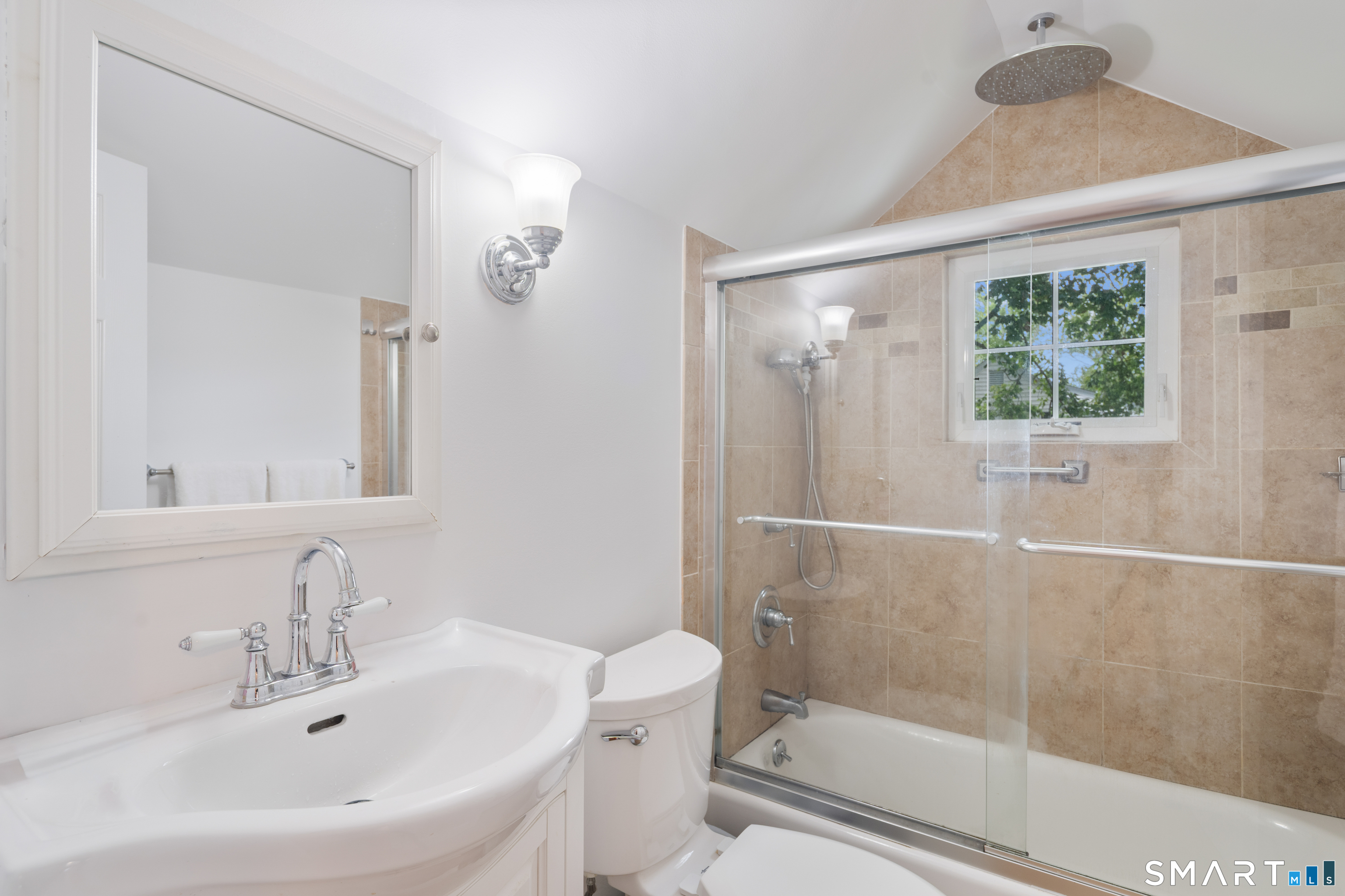 Bathroom with a white vanity and toilet beside a tan-tiled, glass-enclosed tub and shower.