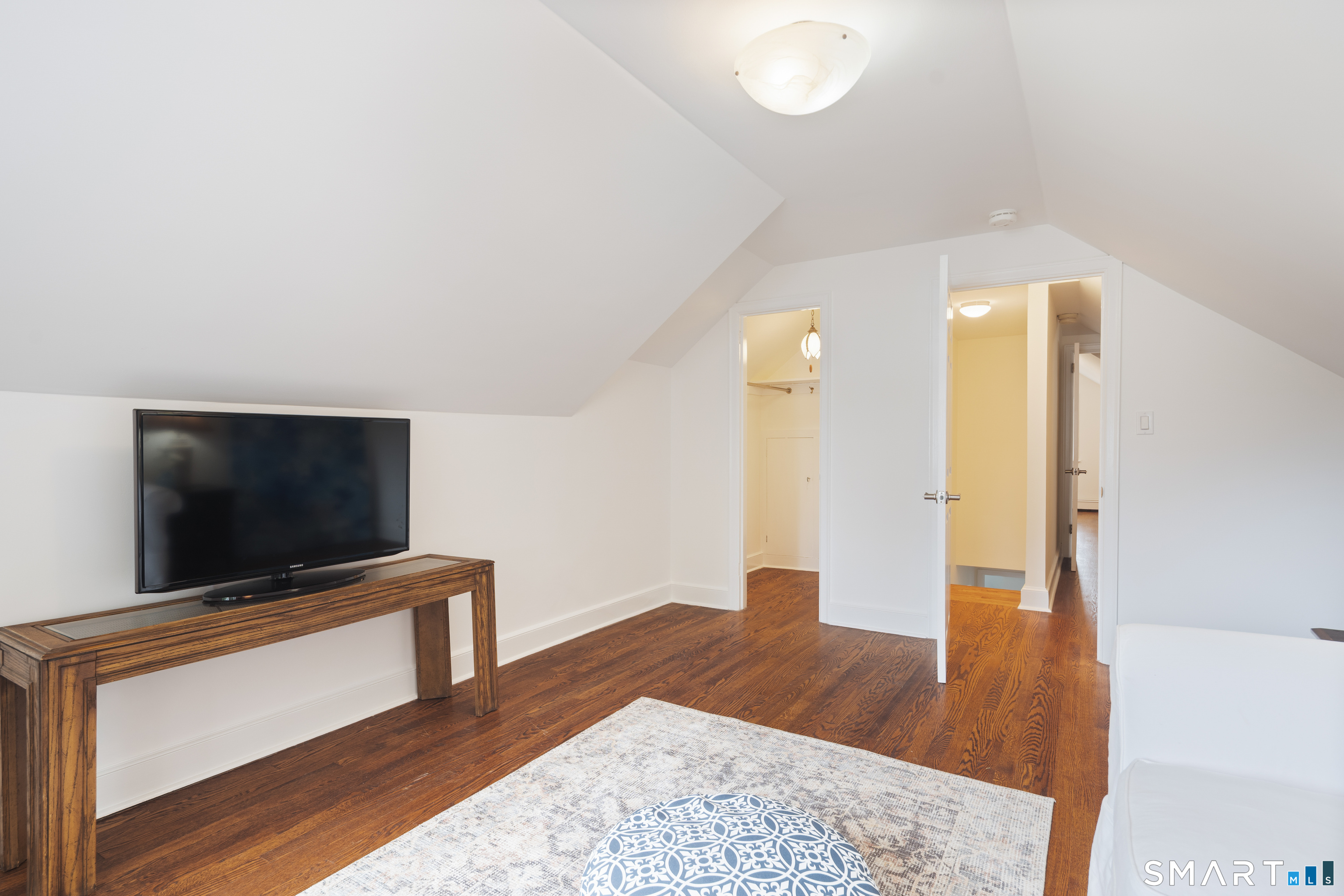 Upper-level sitting room with sloped ceilings, hardwood floors, a television, patterned rug, and white sofa.
