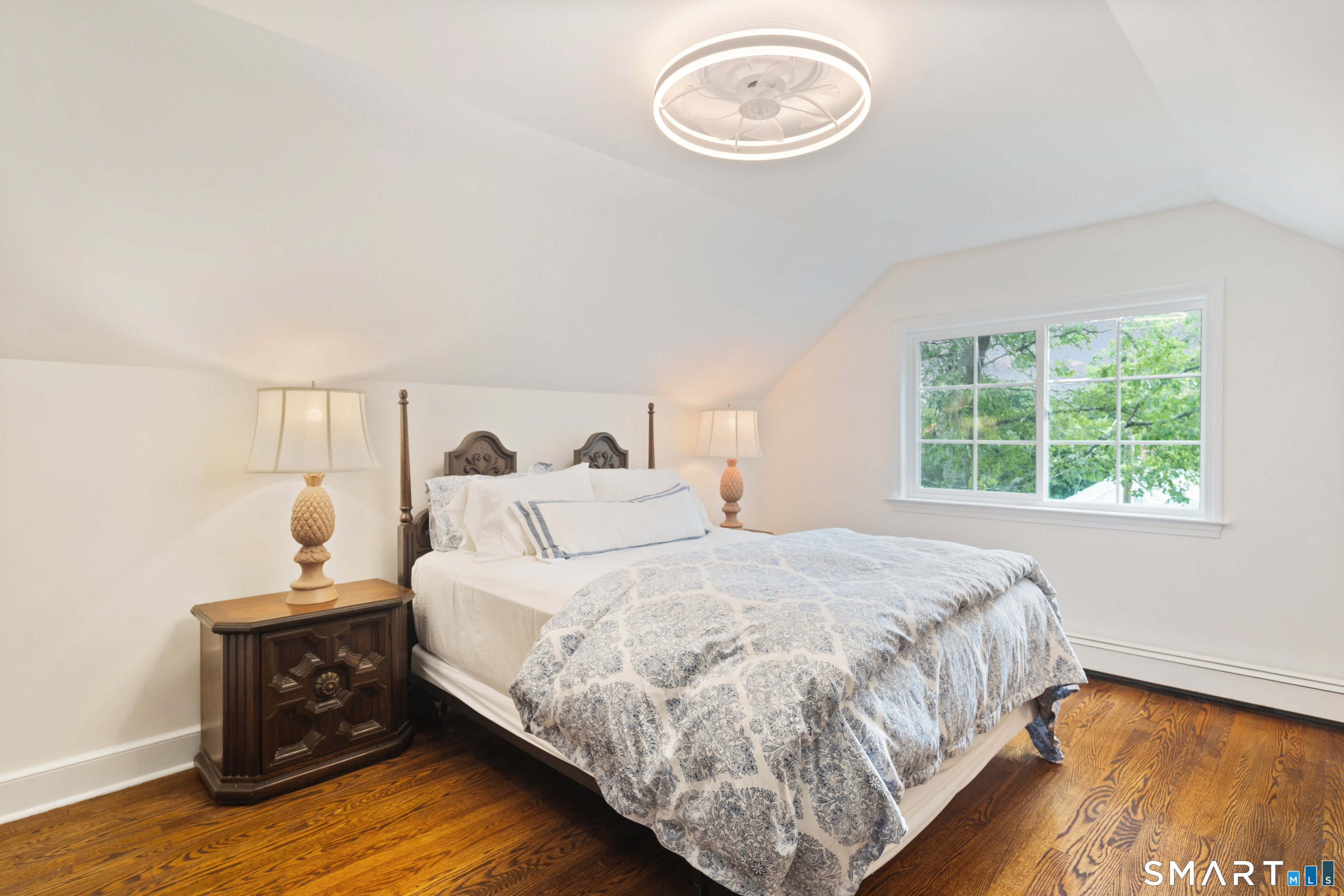 Furnished bedroom with patterned bedding, hardwood floors, and a large window overlooking leafy trees.