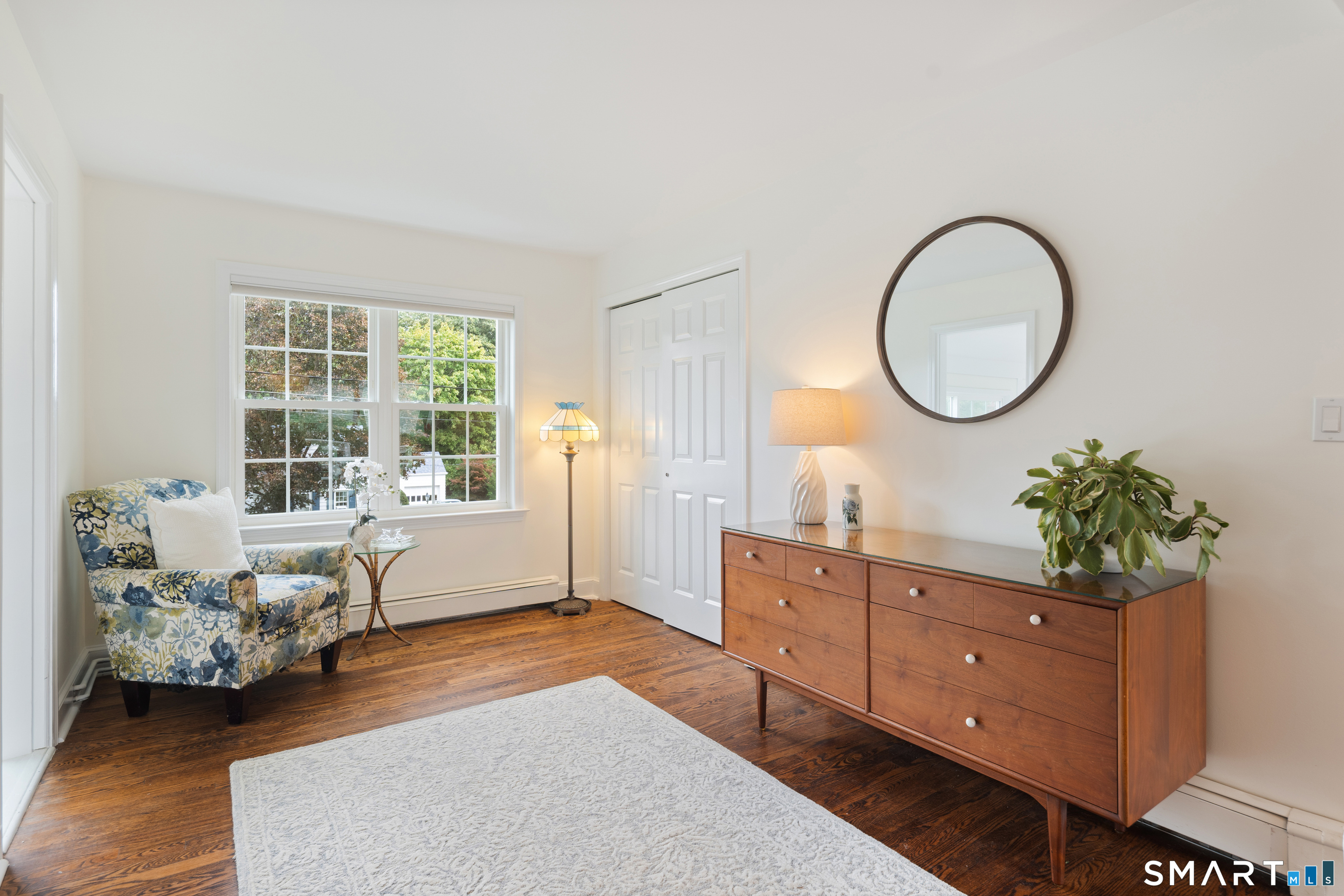Bright sitting room with a patterned armchair, large window, and wooden dresser beneath a round mirror.