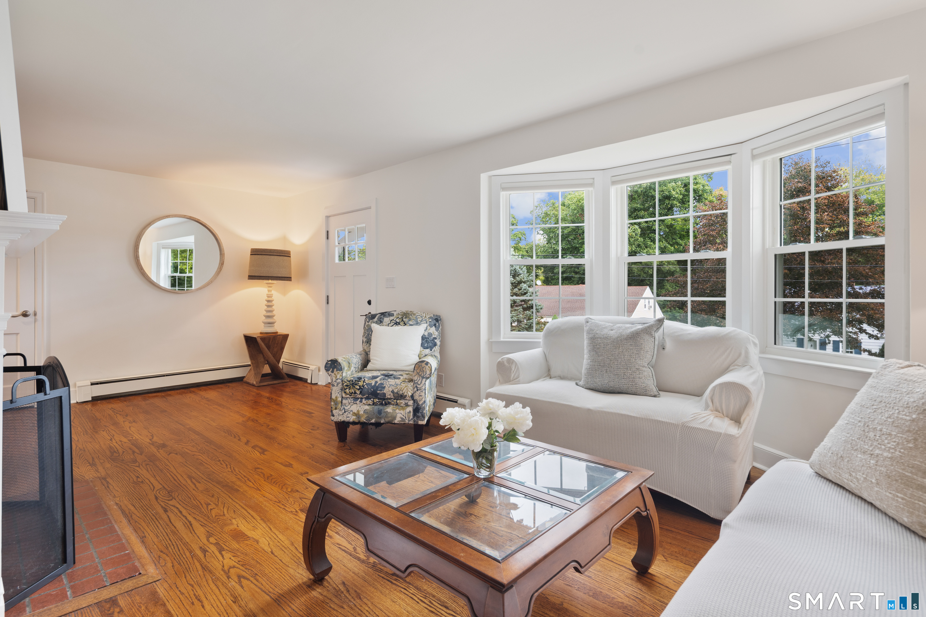 Furnished living room with hardwood floors, a white sofa, and large windows overlooking leafy trees.