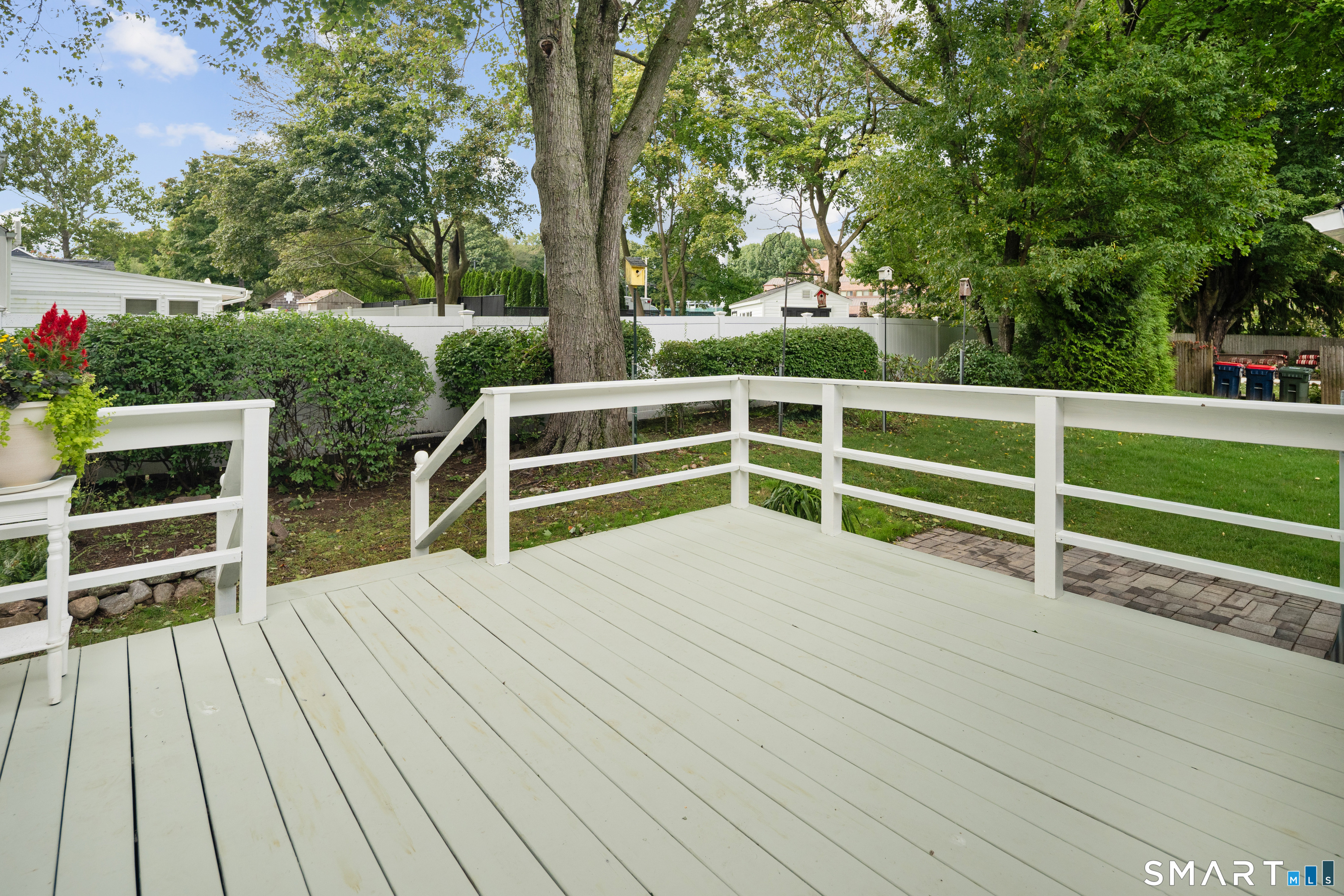 White backyard deck overlooking a tree-lined lawn, with broad railings and a potted flowering plant.