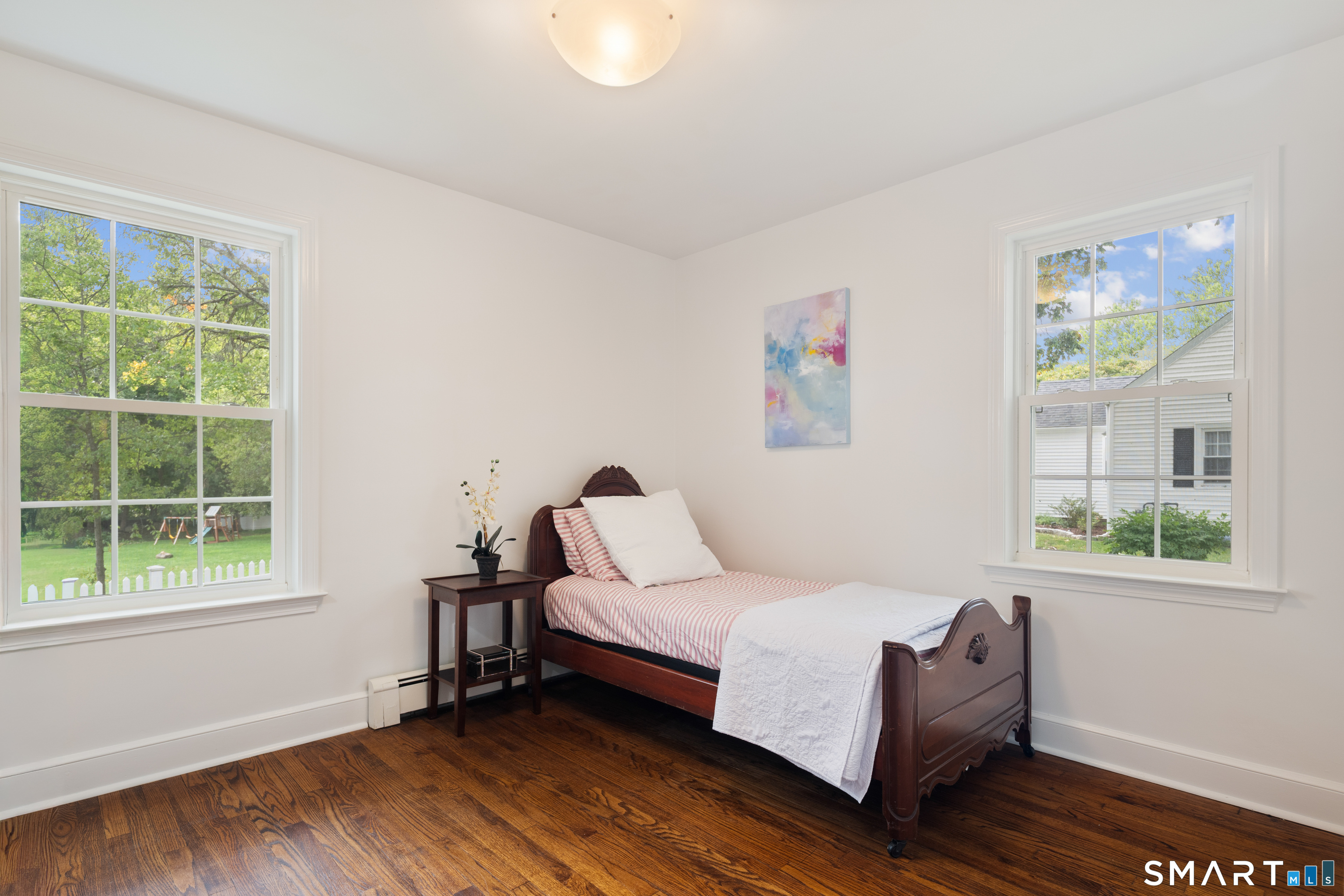 Furnished bedroom with a dark wood single bed, striped bedding, and two large windows overlooking a green yard.