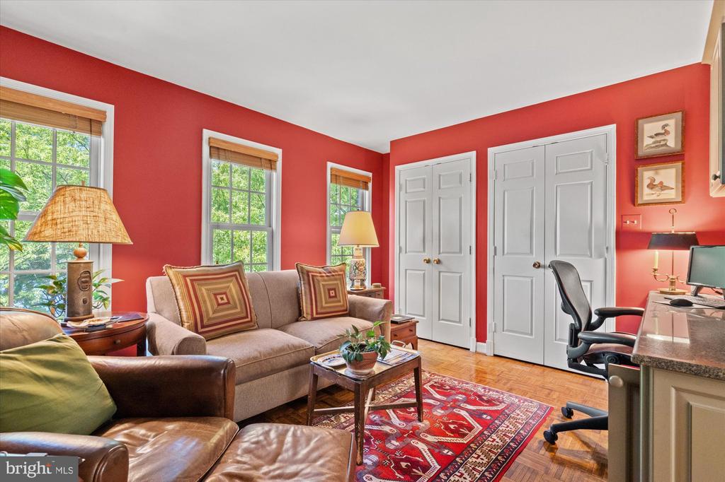 Furnished red-walled room with a sitting area, office desk, parquet floors, and windows overlooking greenery.