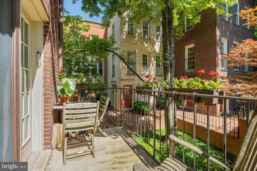 Private patio with wooden chairs and potted plants overlooking leafy brick townhouses.