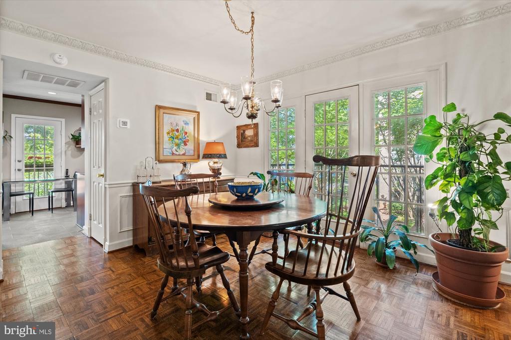 Dining room with a round wooden table, parquet floors, chandelier, and tall windows overlooking greenery.