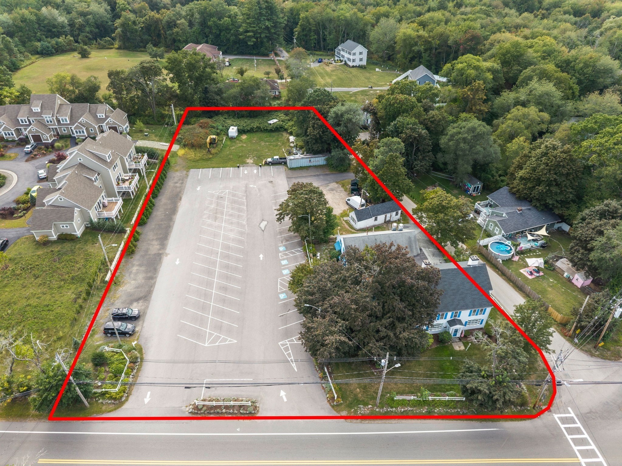 Aerial view of a roadside property outlined in red, with a large parking lot and a tree-shaded building.
