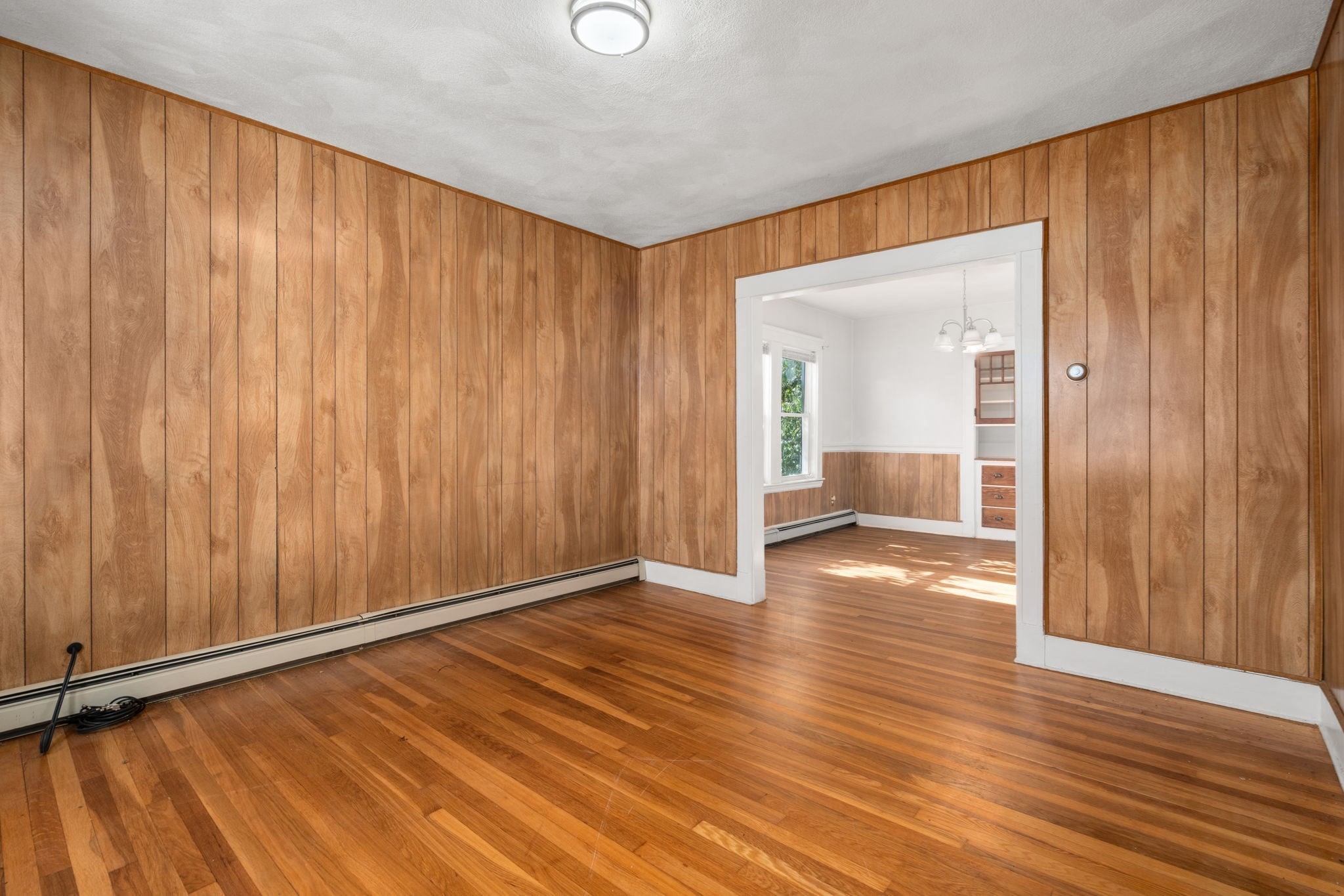 Empty wood-paneled room with hardwood floors and a wide doorway to a bright adjoining dining area.