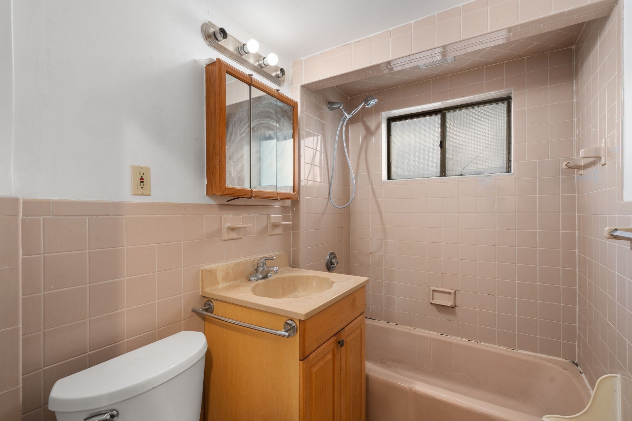 Bathroom with pink tile, a bathtub and shower, and a wood vanity beneath a mirrored cabinet.