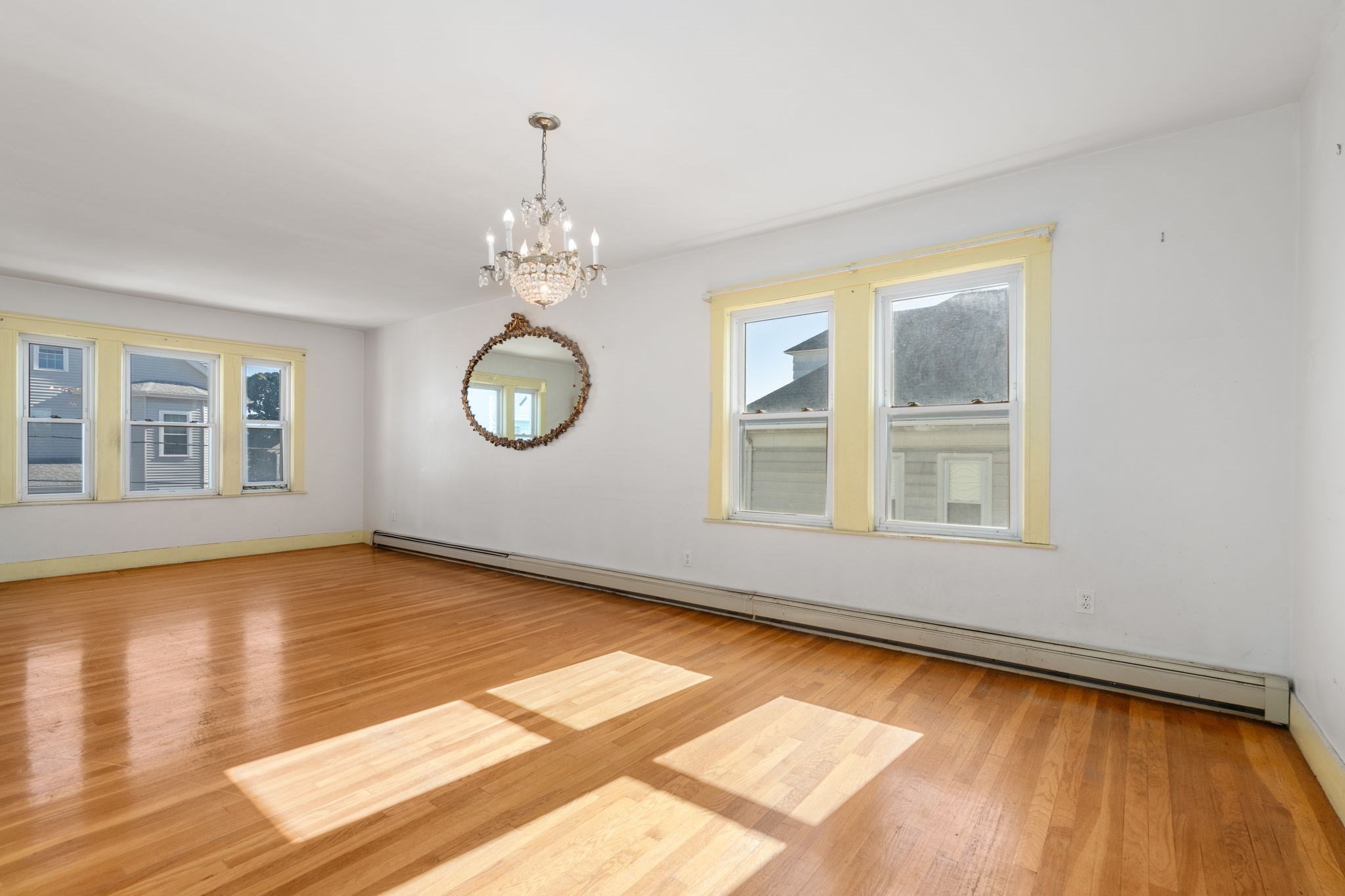 Vacant living room with hardwood floors, yellow-trimmed windows, a chandelier, and an ornate round mirror.
