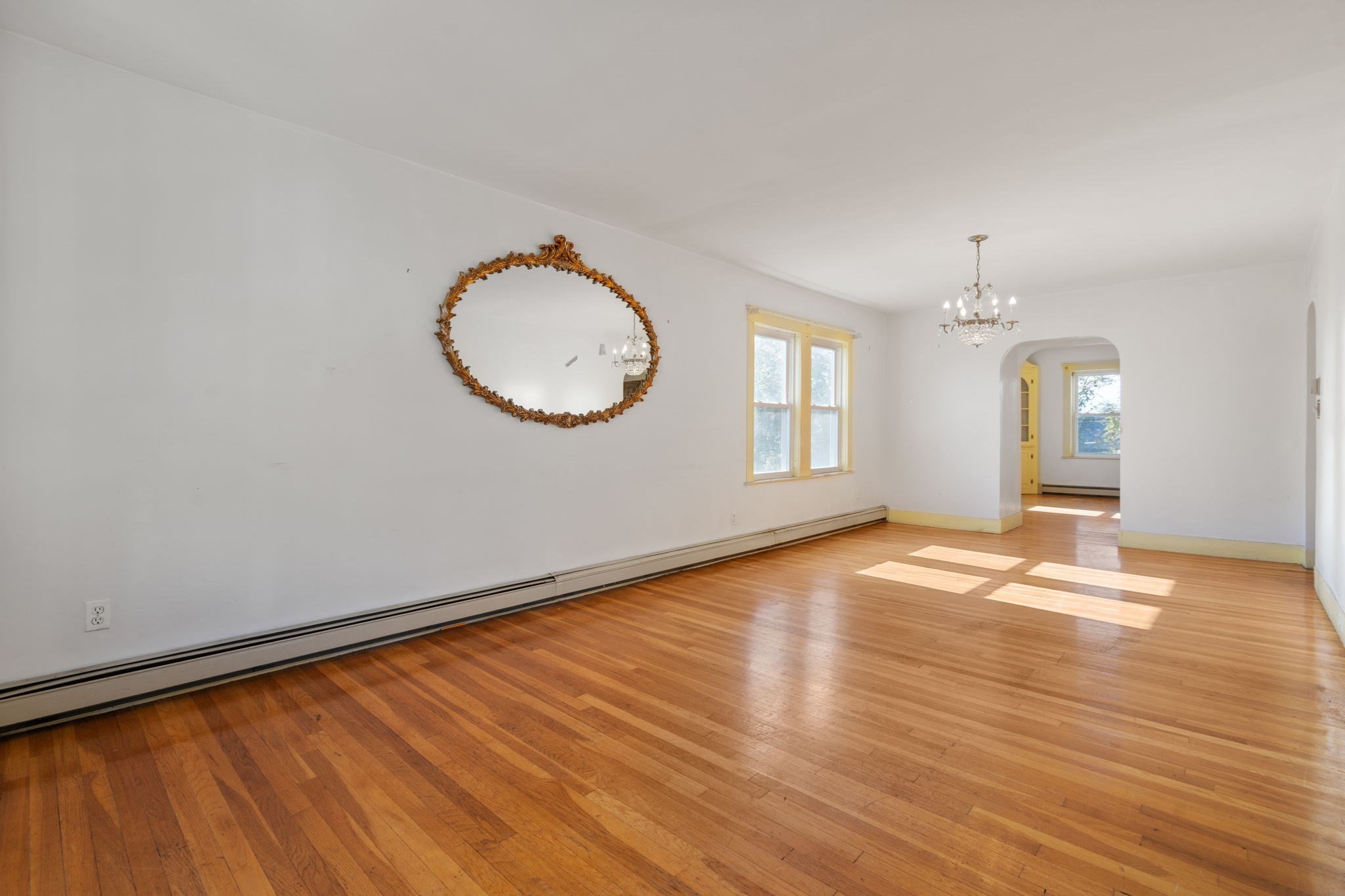 Vacant living room with polished hardwood floors, an ornate oval mirror, and an arched doorway.