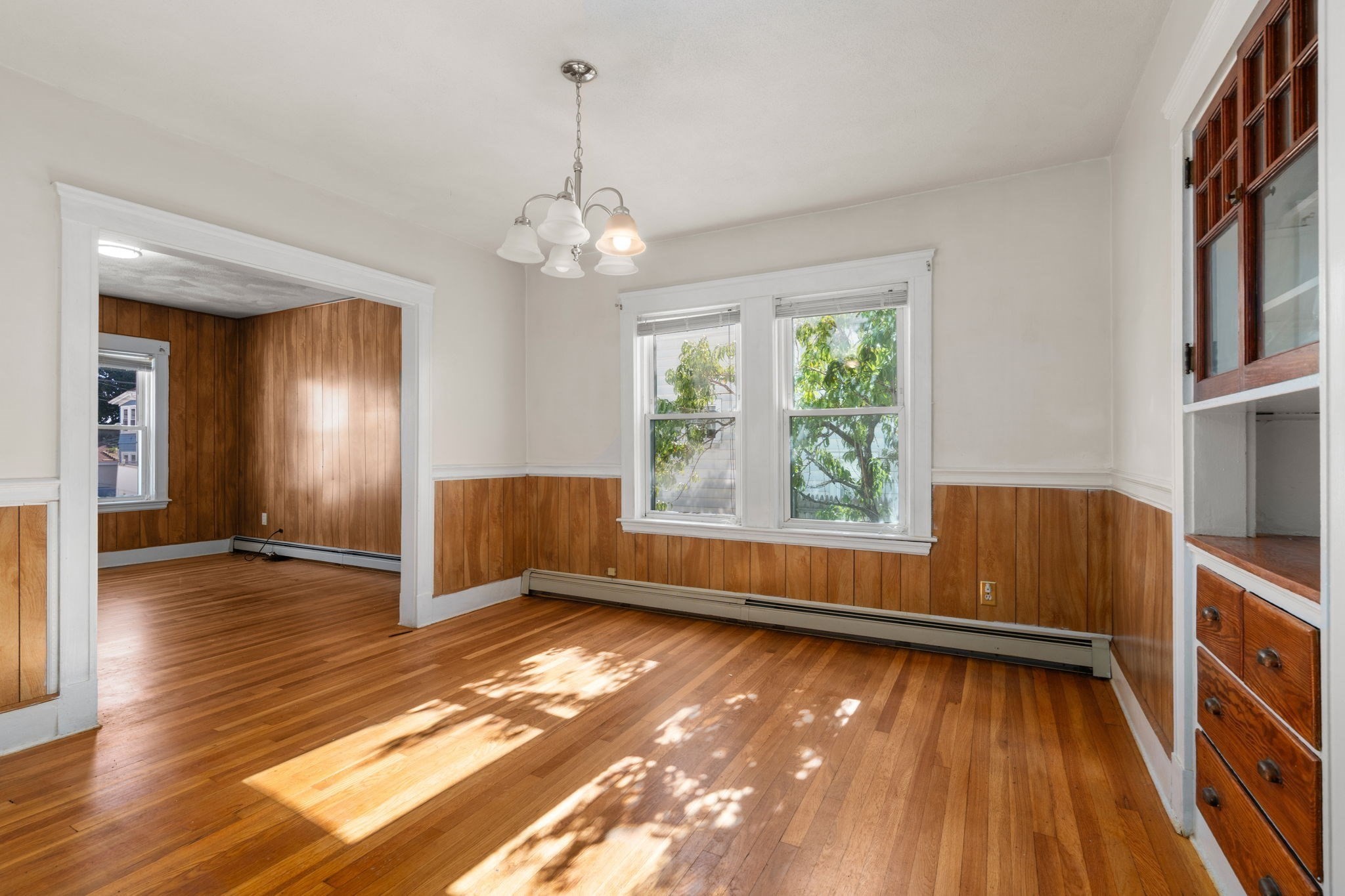 Vacant room with hardwood floors, wood-paneled walls, built-in cabinets, and sunlight through two windows.