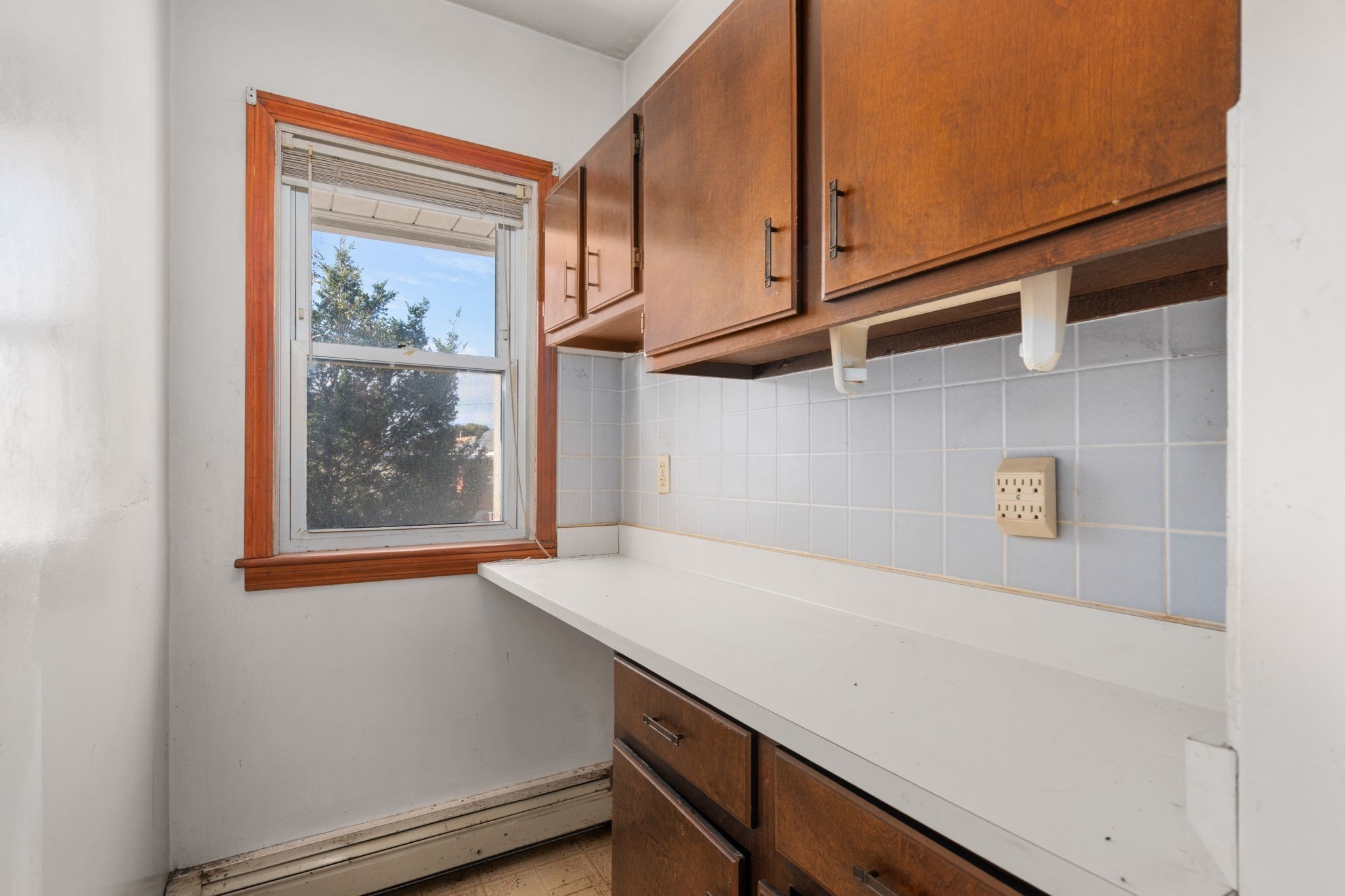 Kitchen with long white countertop, dark wood cabinets, pale blue tile backsplash, and a window overlooking trees.