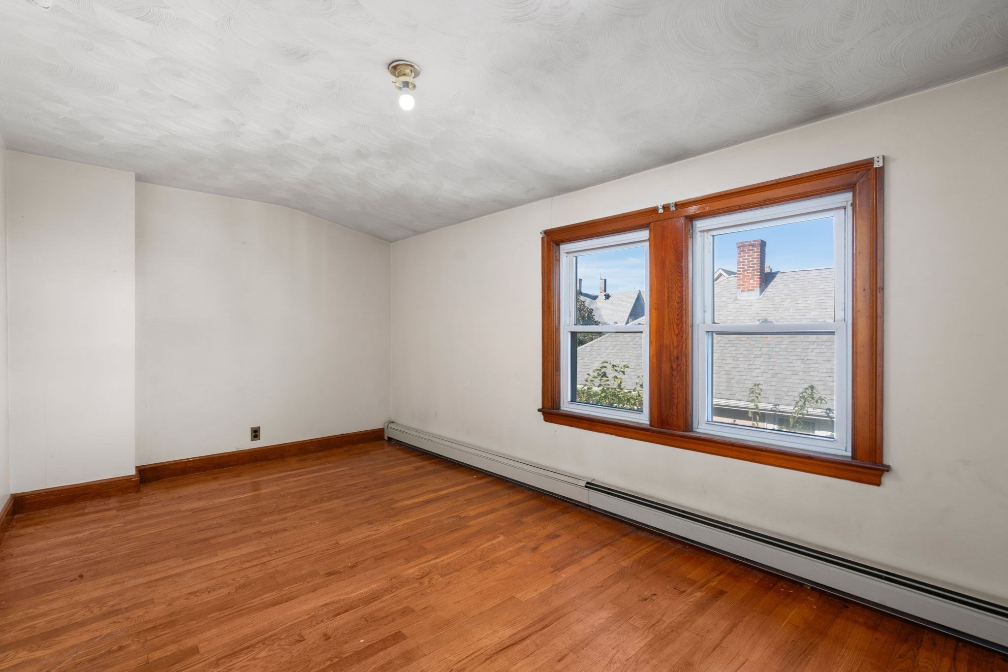 Empty room with hardwood floors, two wood-trimmed windows, and a baseboard heater.