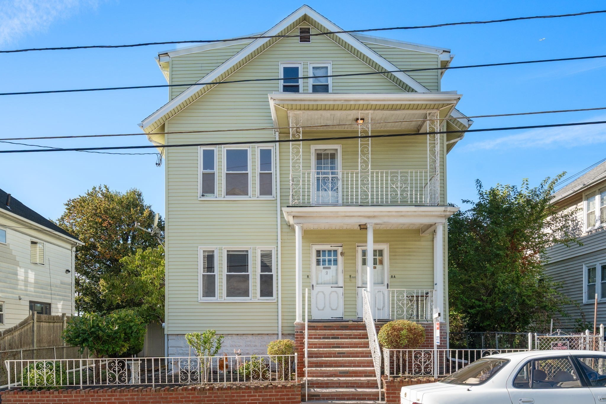 Pale green multi-story house with ornate white balconies, brick steps, and a fenced front yard.