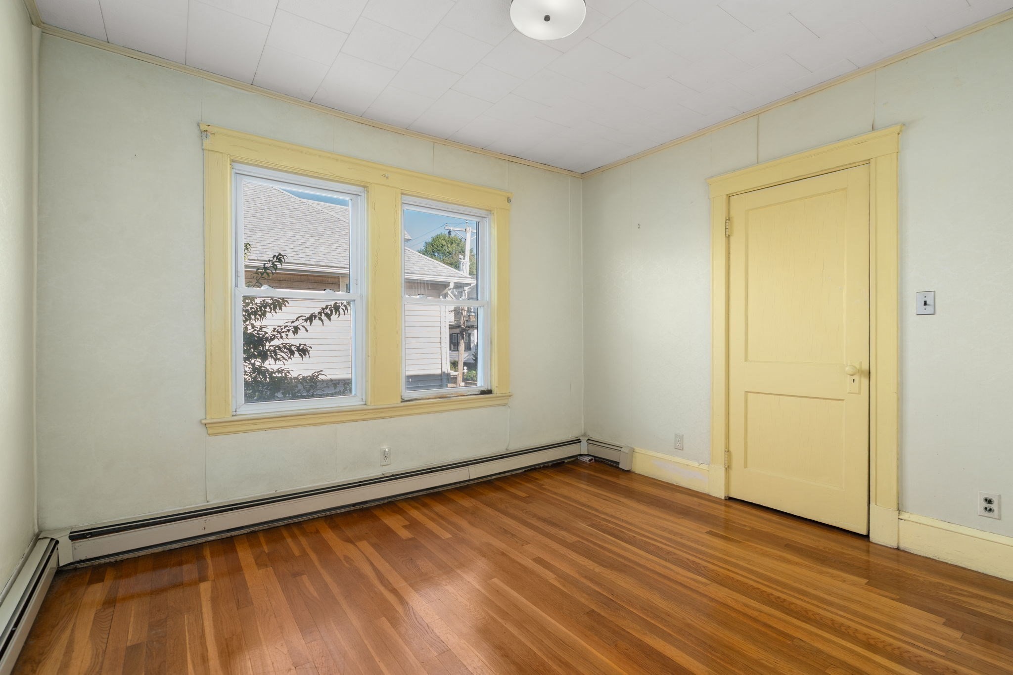 Empty room with polished hardwood floors, yellow trim, and two windows.