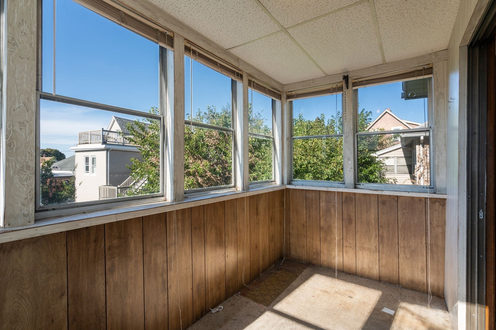 Empty sunroom with wraparound windows, wood-paneled walls, and sunlight across the carpet.