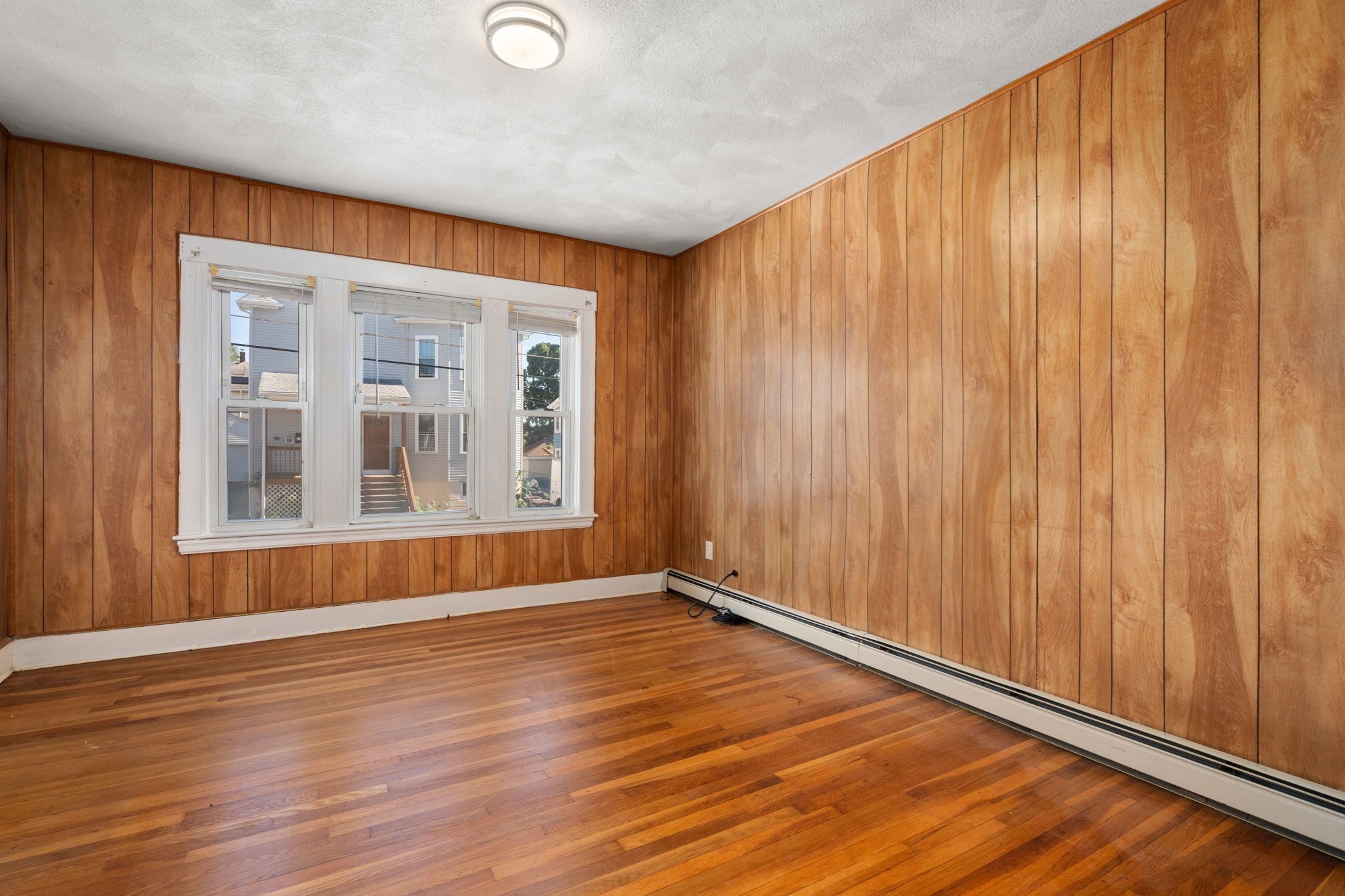 Empty room with wood-paneled walls, hardwood floors, and a wide three-section window.