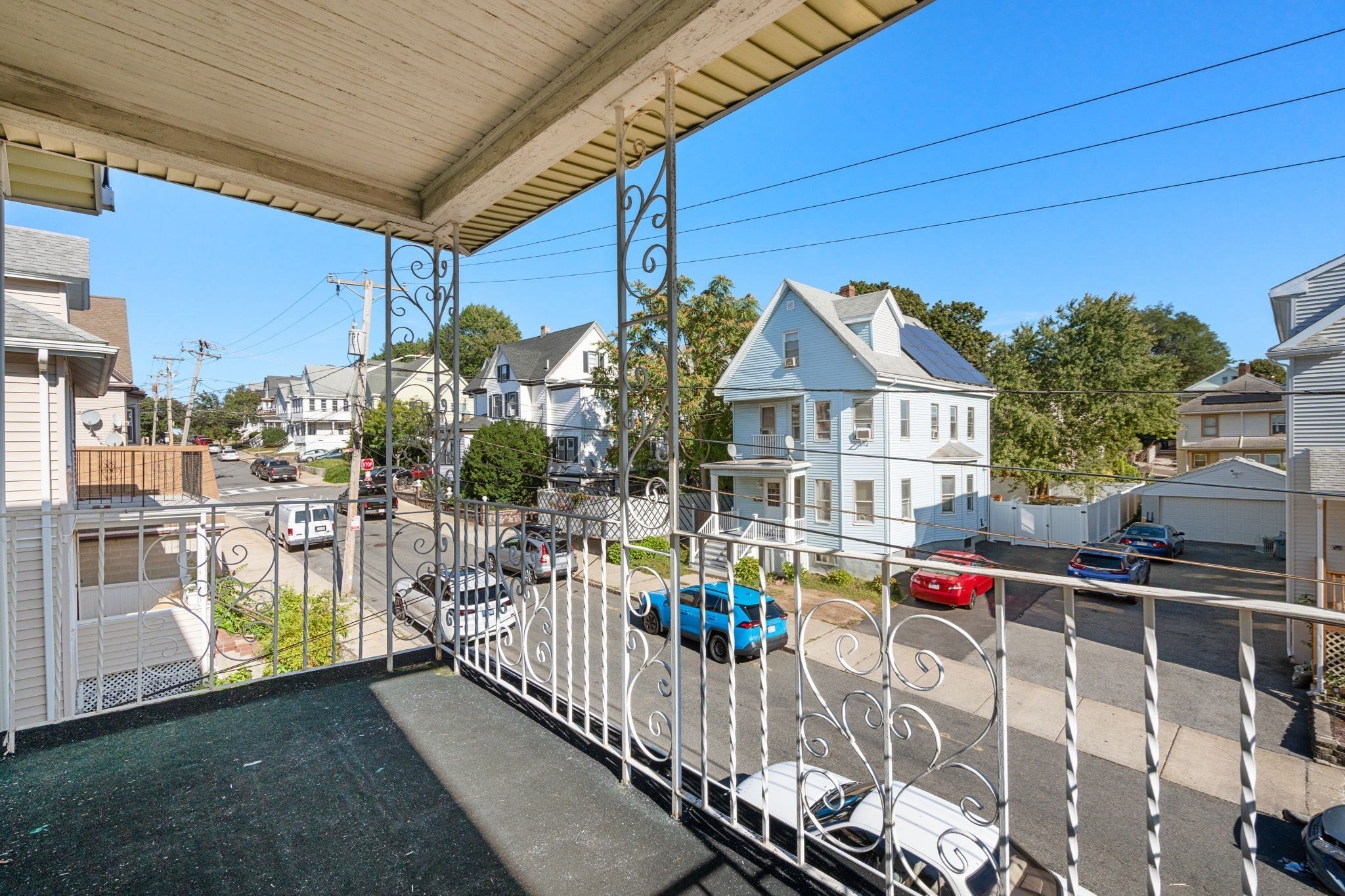 Covered porch with ornate metal railings overlooking a residential street and neighboring homes.