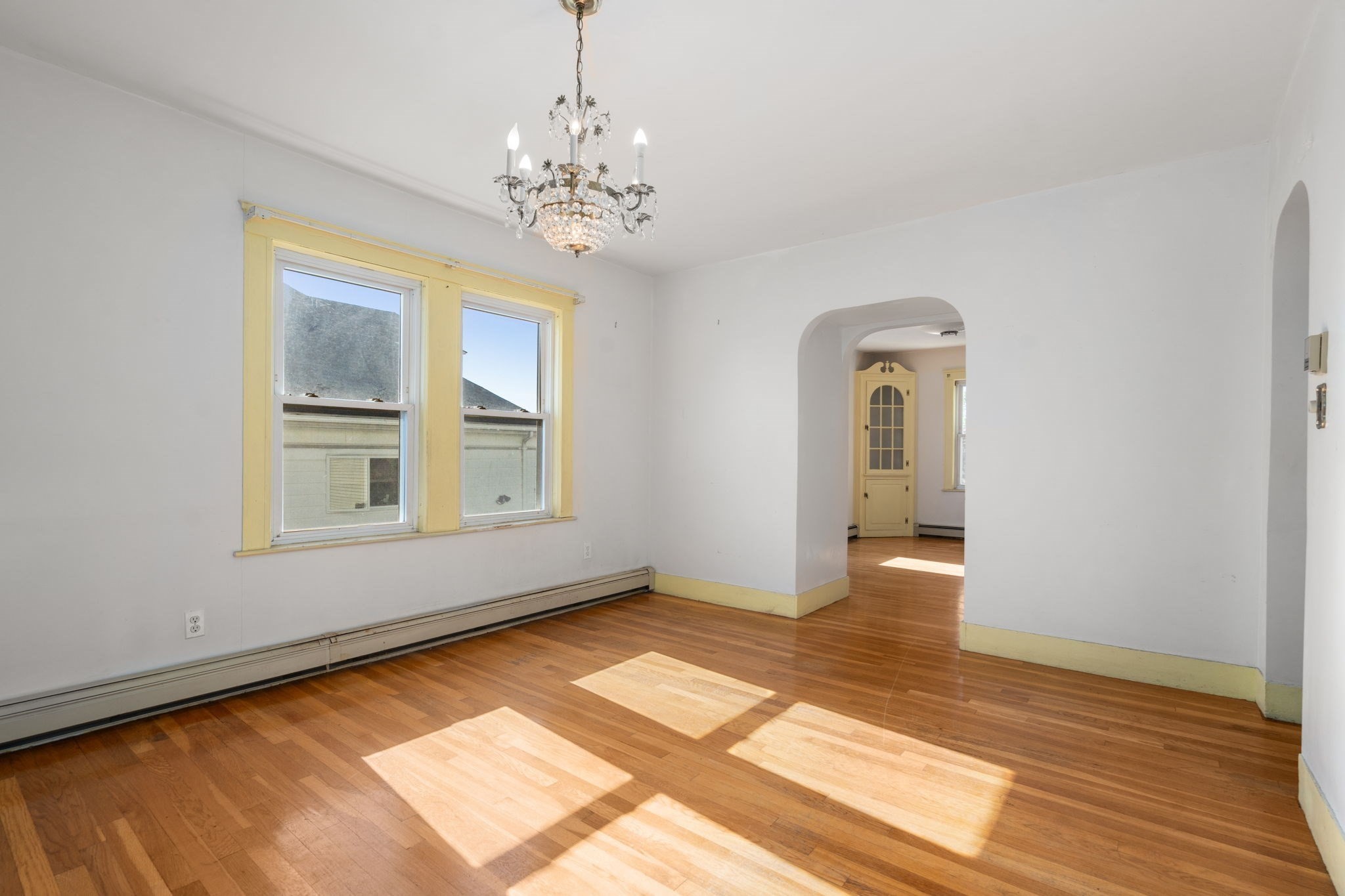 Empty room with hardwood floors, yellow-trimmed windows, a chandelier, and an arched doorway.
