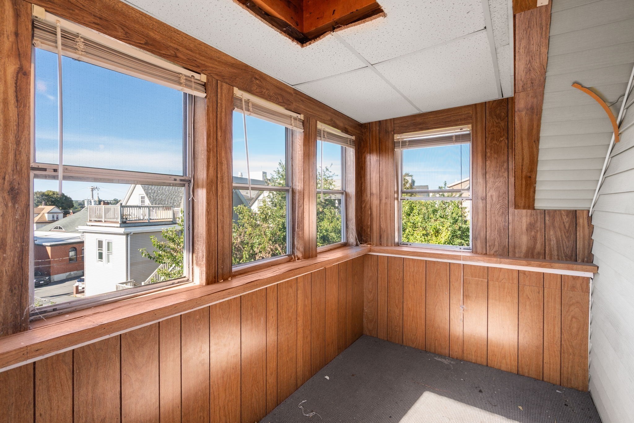 Enclosed sunroom with wood paneling, broad window ledges, and views of neighboring homes and trees.