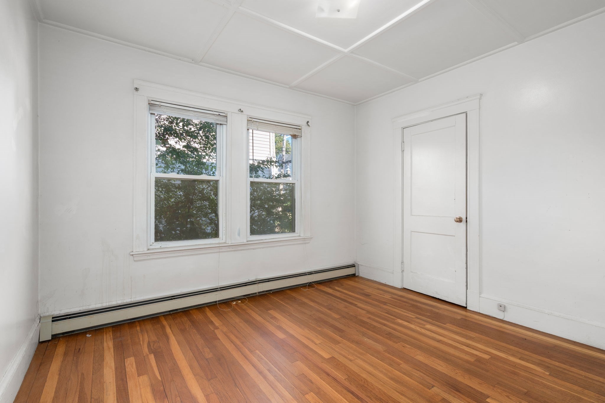 Empty room with hardwood floors, two windows, and a long baseboard heater.