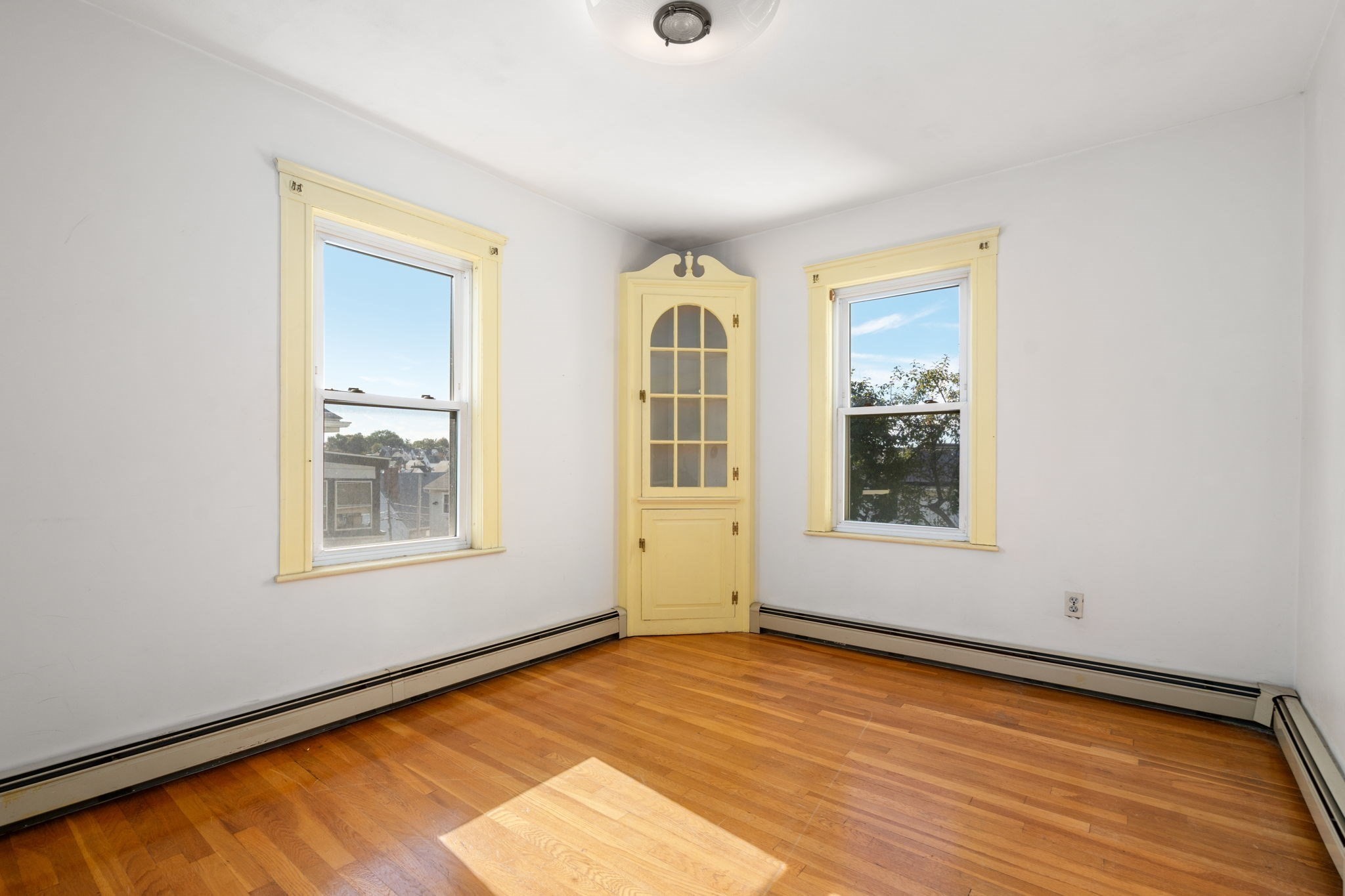 Empty room with hardwood floors, pale yellow window trim, and a matching built-in corner cabinet.