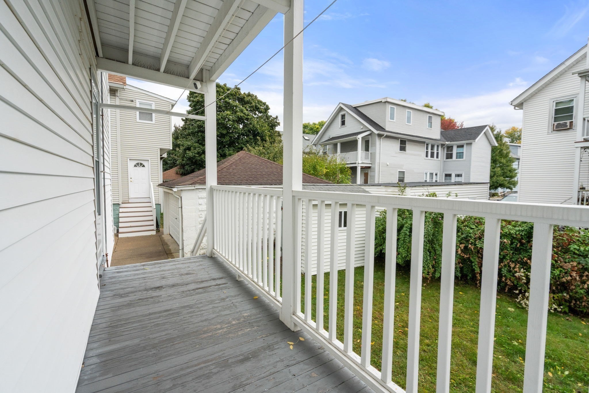Covered porch with gray decking and white railings overlooking a lawn and neighboring houses.