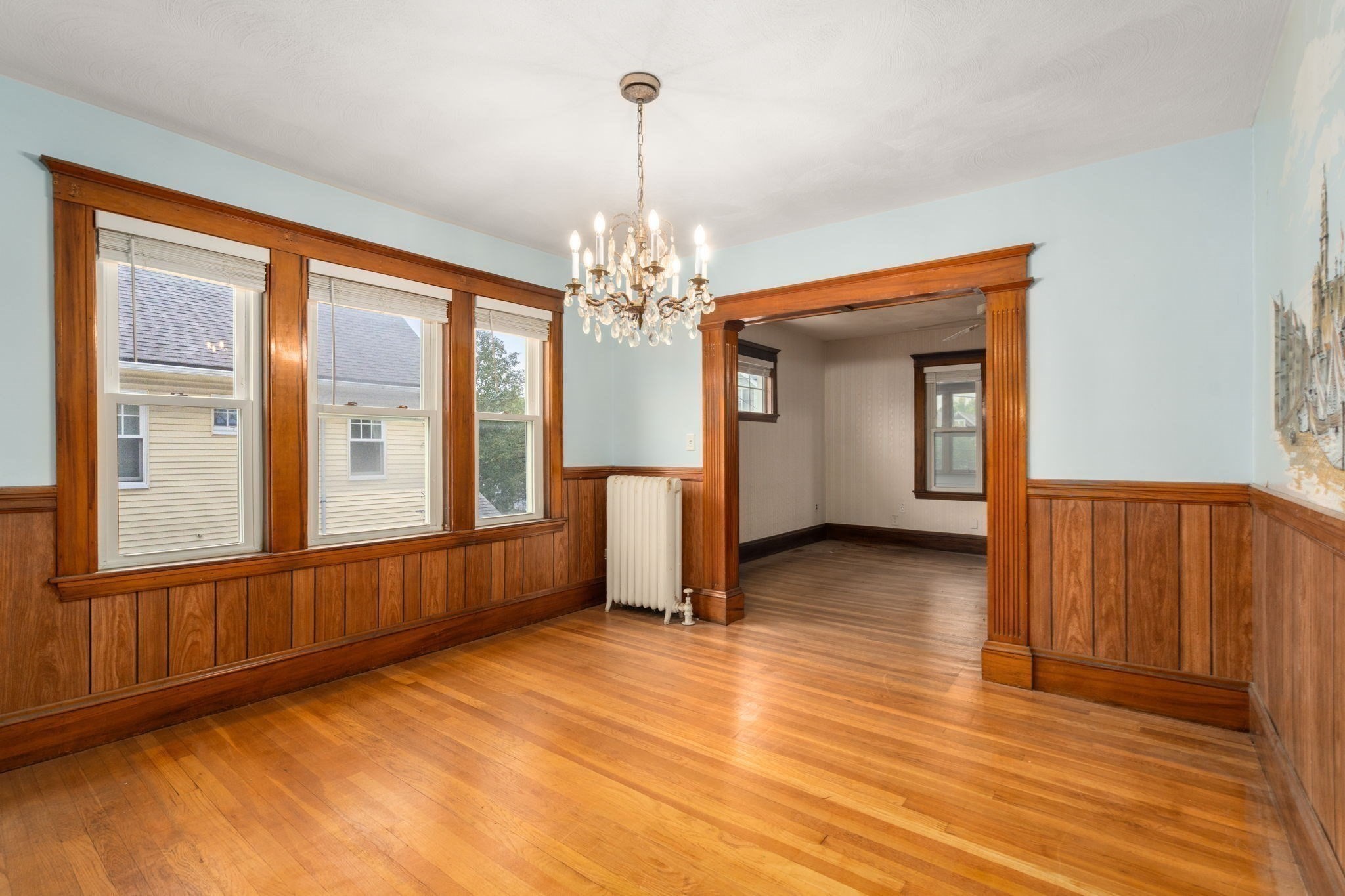 Empty room with hardwood floors, wood-paneled walls, tall windows, and a chandelier.