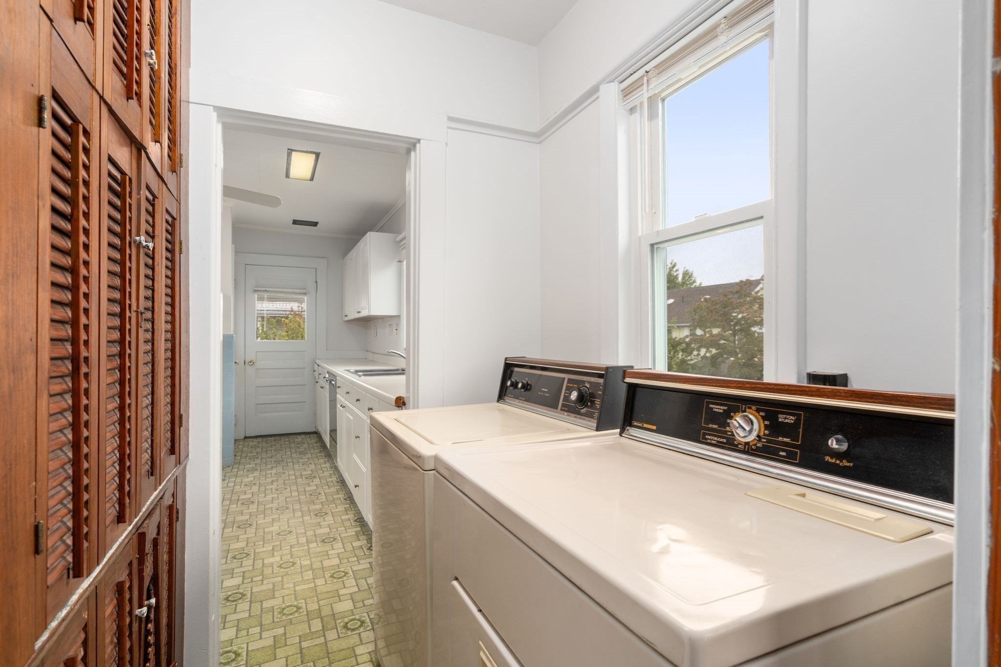 Laundry room with two white top-loading machines, white cabinetry, patterned flooring, and a large window.