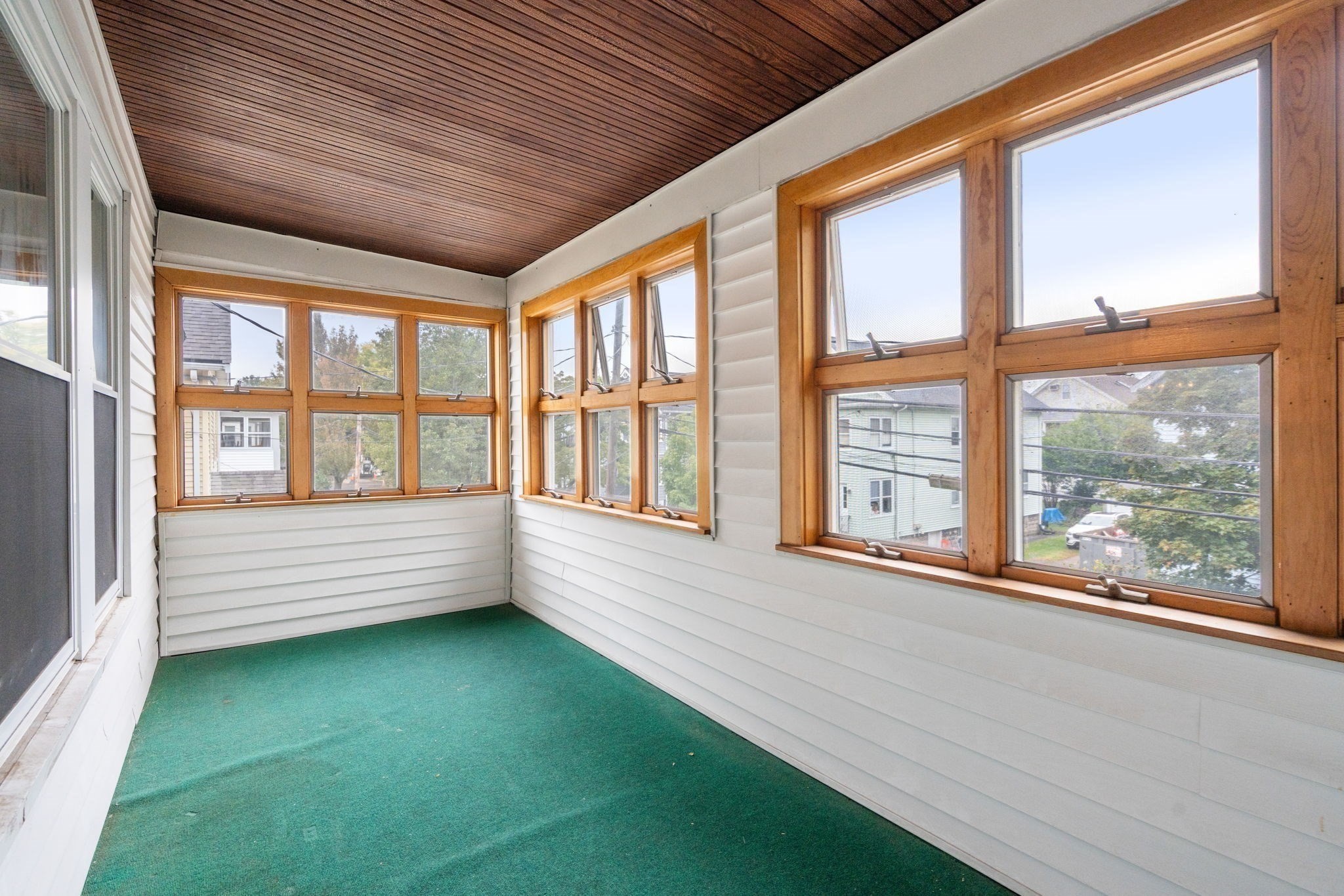 Vacant sunroom with green carpet, a wood-plank ceiling, and rows of wood-framed windows.