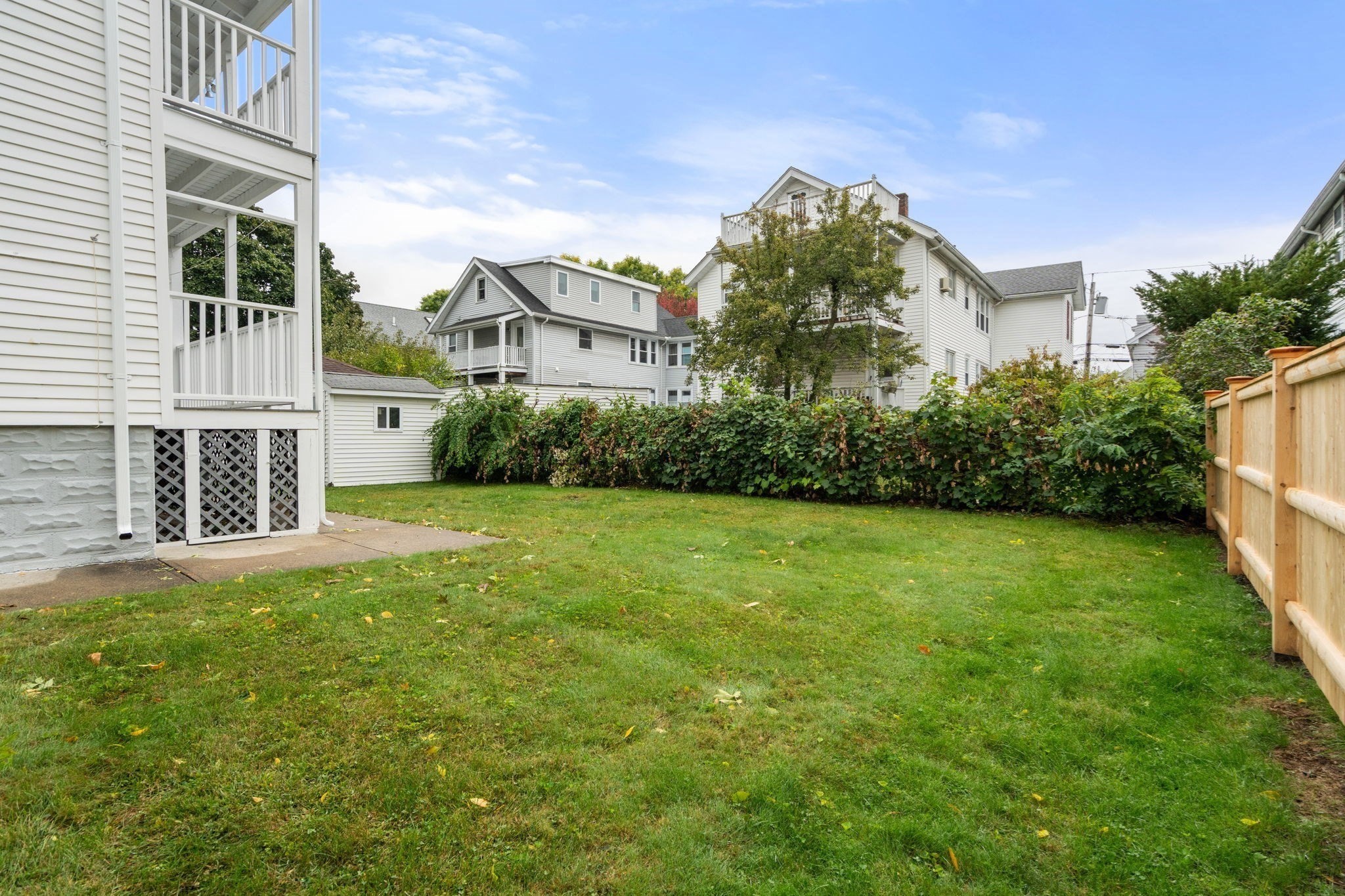 Backyard lawn bordered by a leafy hedge and wooden fence, with raised porches along the house.