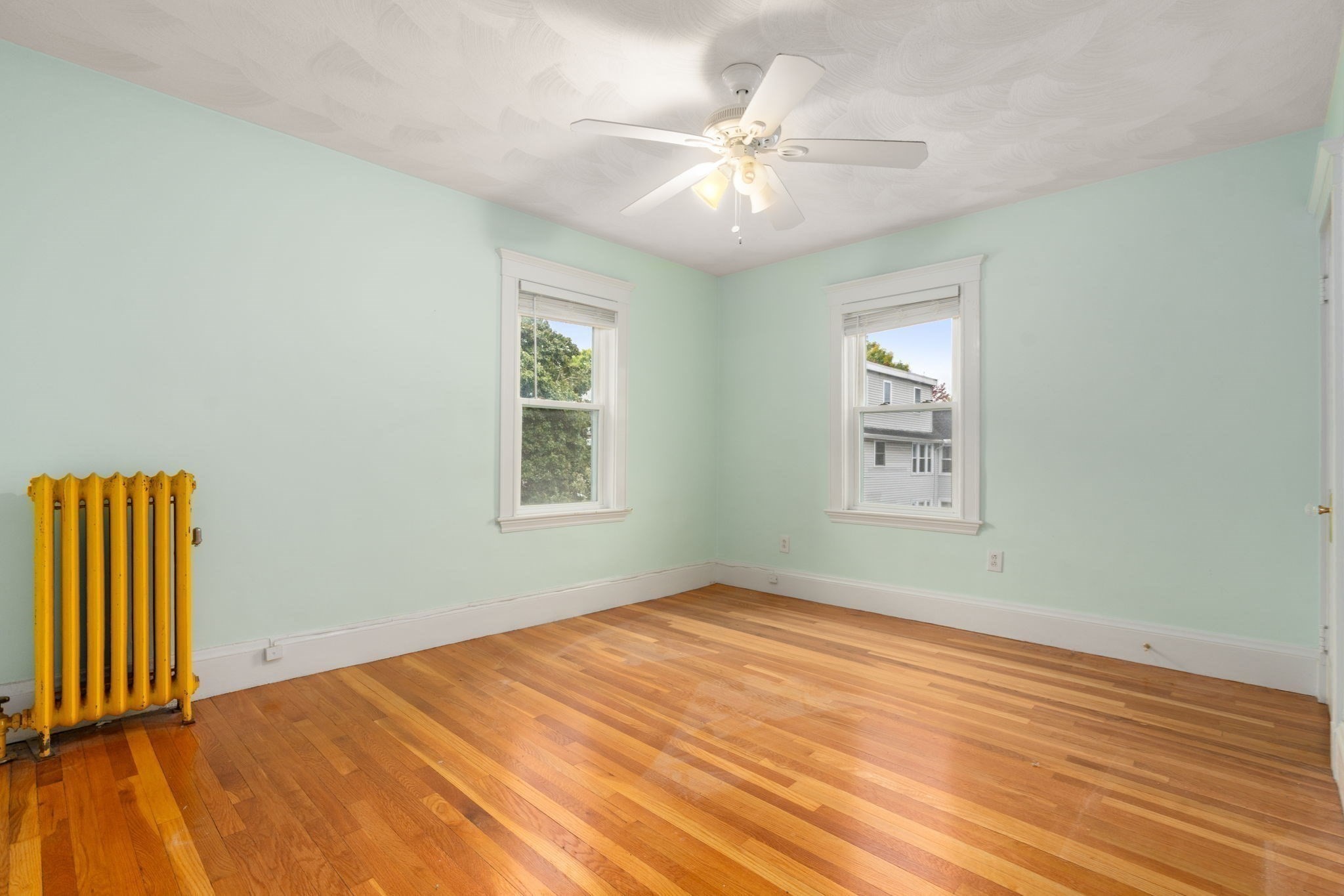 Empty room with mint-green walls, hardwood floors, two windows, and a yellow radiator.