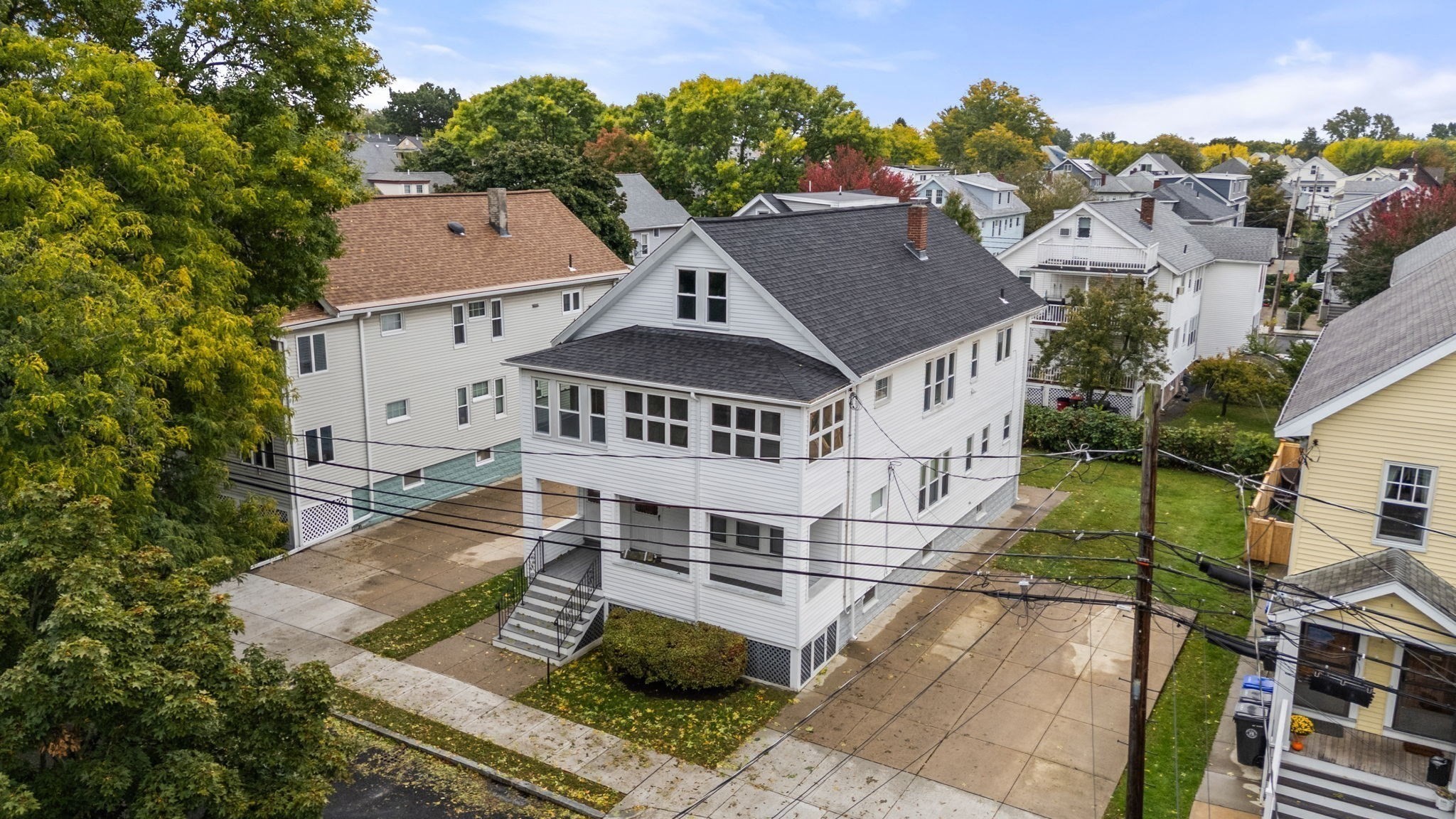 White multi-story home with a dark gabled roof and enclosed porch, viewed from above among autumn trees.