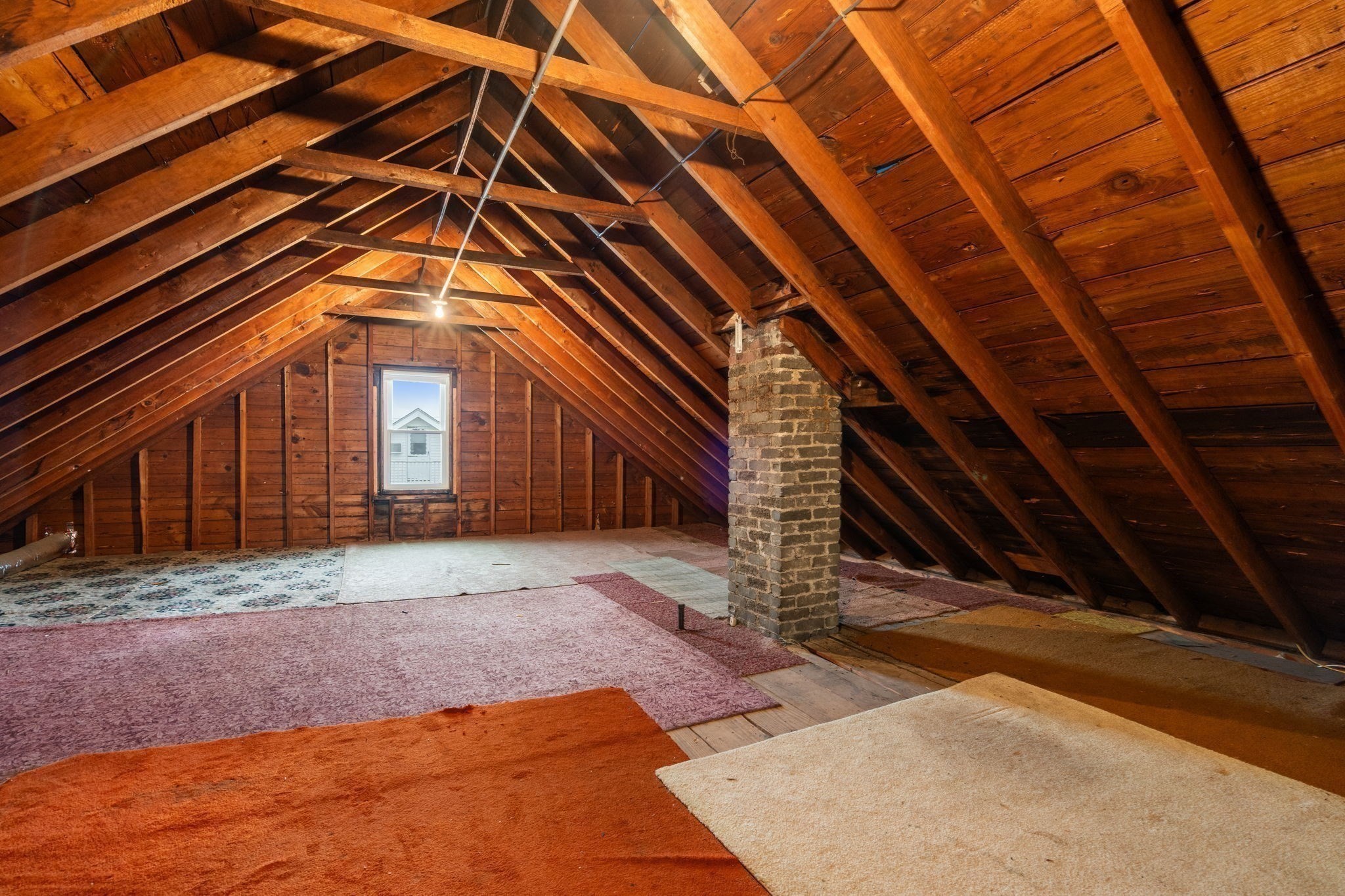 Unfinished attic with exposed timber rafters, a brick chimney, and carpet remnants.