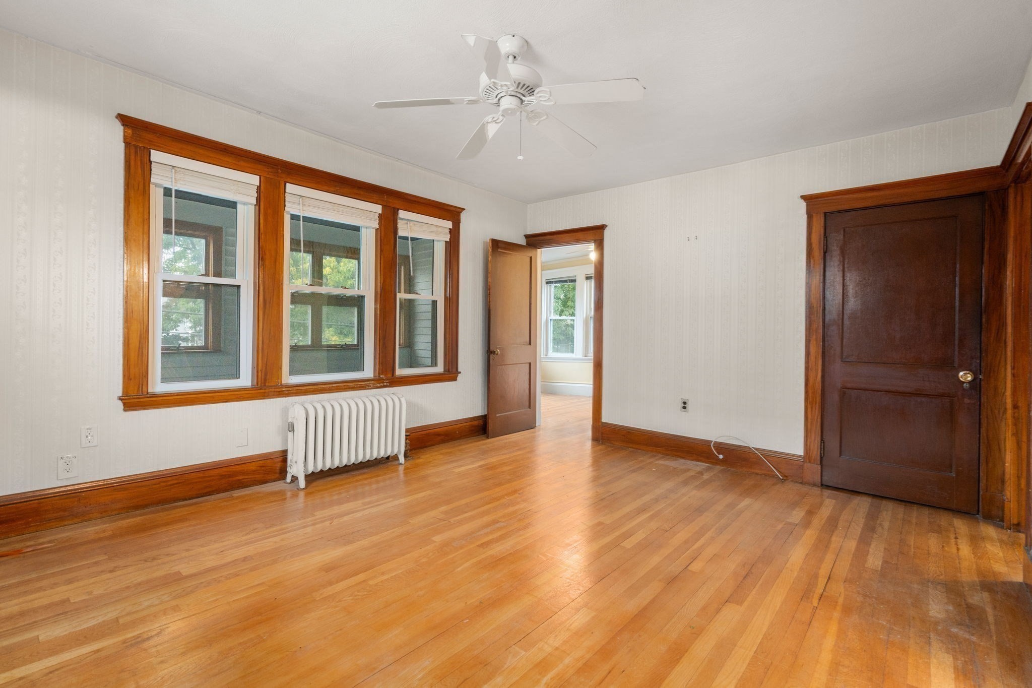 Empty room with polished hardwood floors, dark wood trim, three windows, and a white radiator.