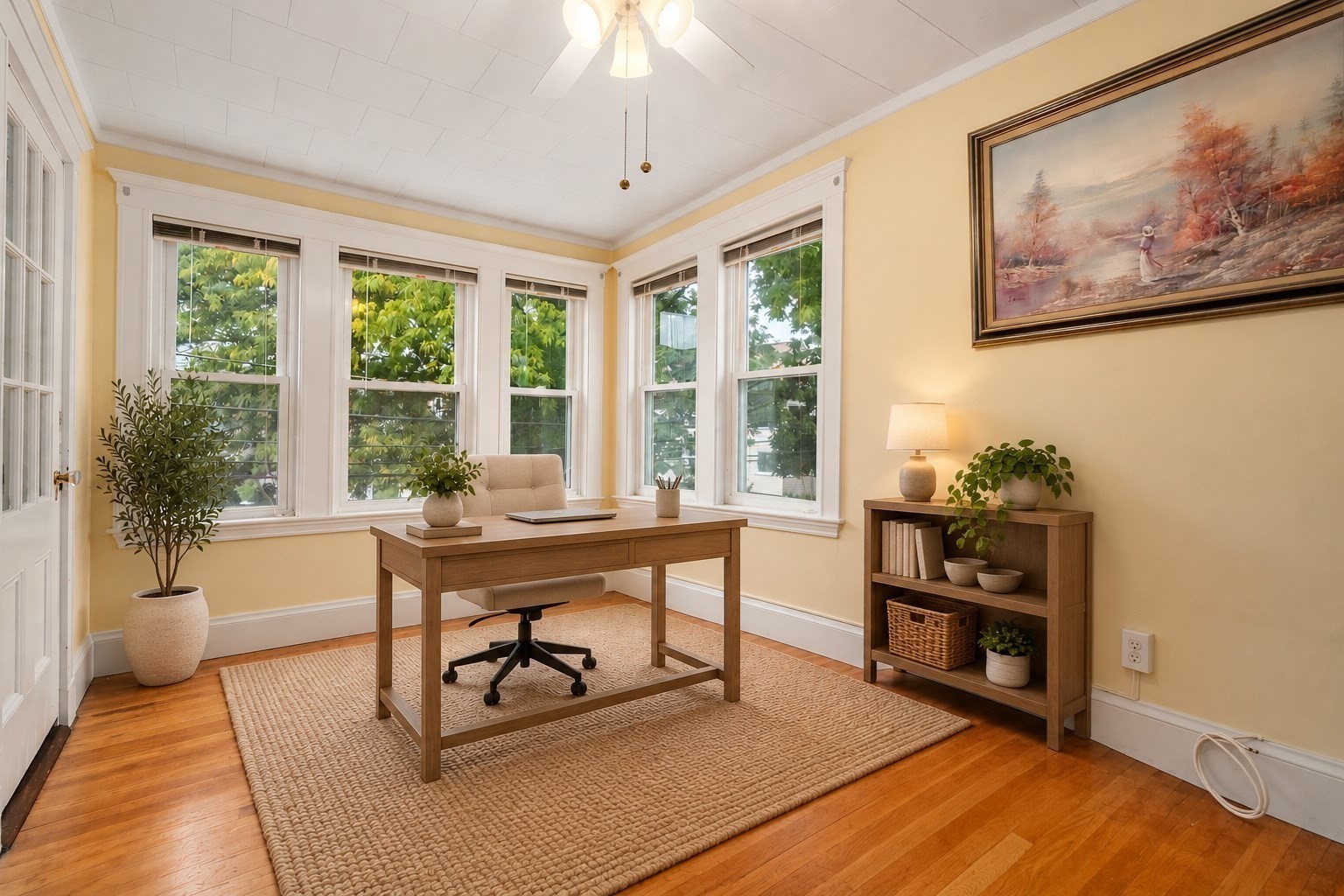 Home office with a wooden desk, woven rug, and large windows overlooking leafy trees.
