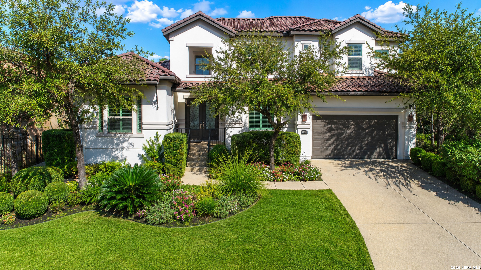 Two-story stucco home with a red tile roof, shaded entry, attached garage, and lush landscaped front yard.
