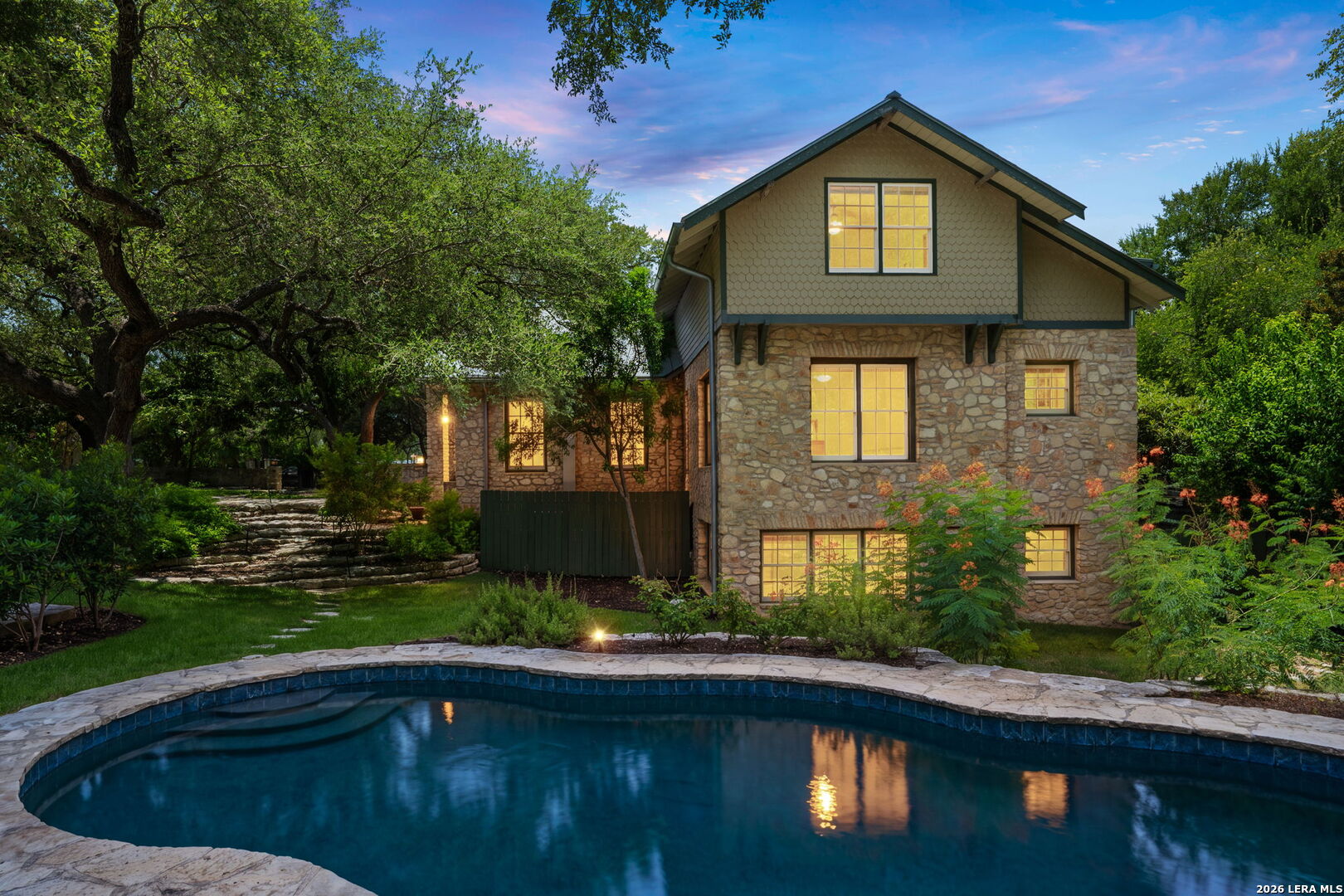 Stone-and-shingle house with a swimming pool and mature trees at dusk.