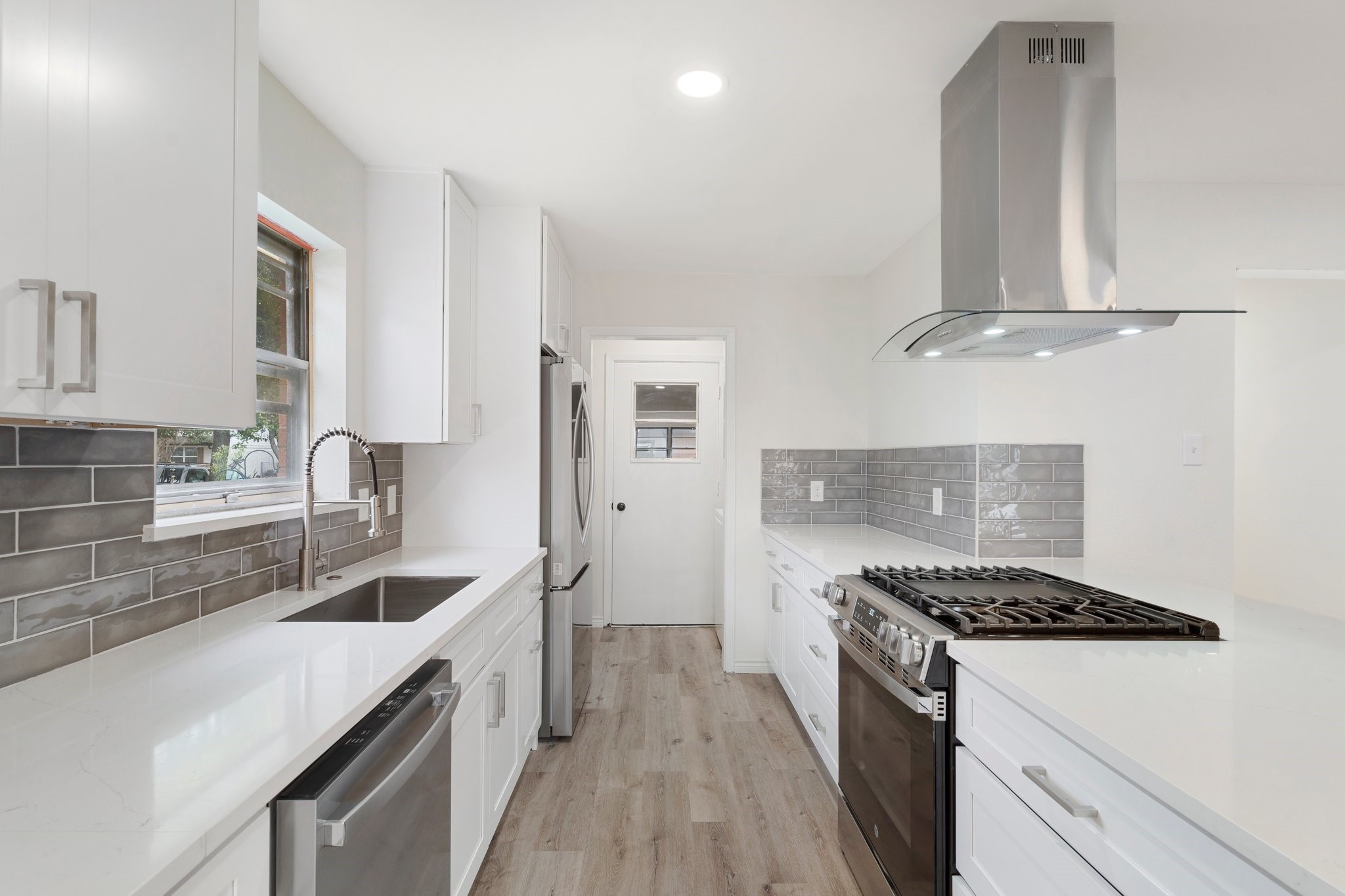 Galley kitchen with white cabinets, gray tile backsplash, stainless steel gas range, and light wood floors.