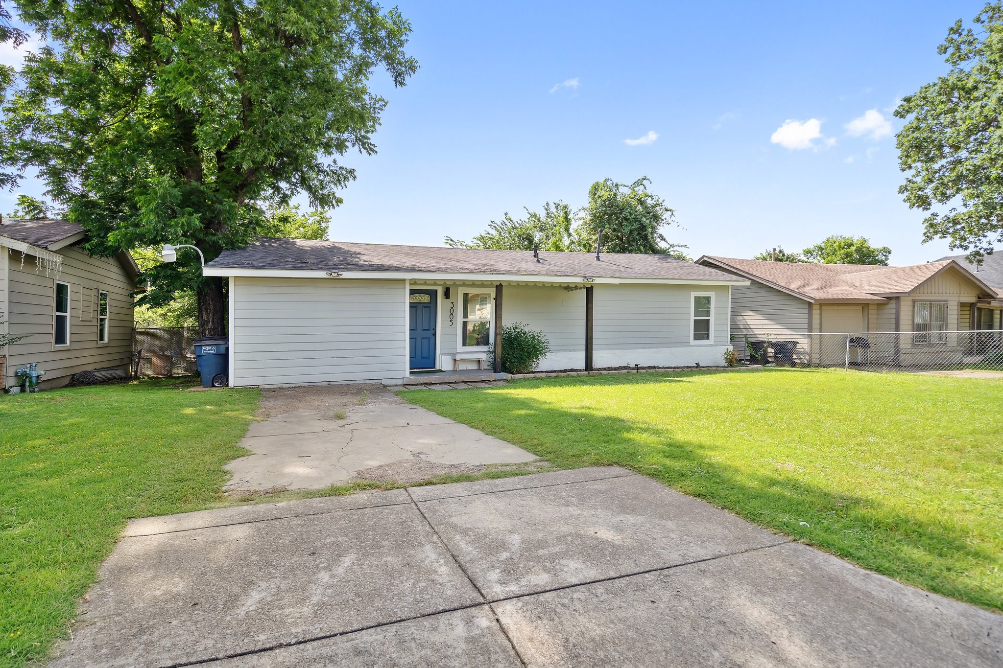 Single-story gray house with a blue front door, broad driveway, and mature trees.