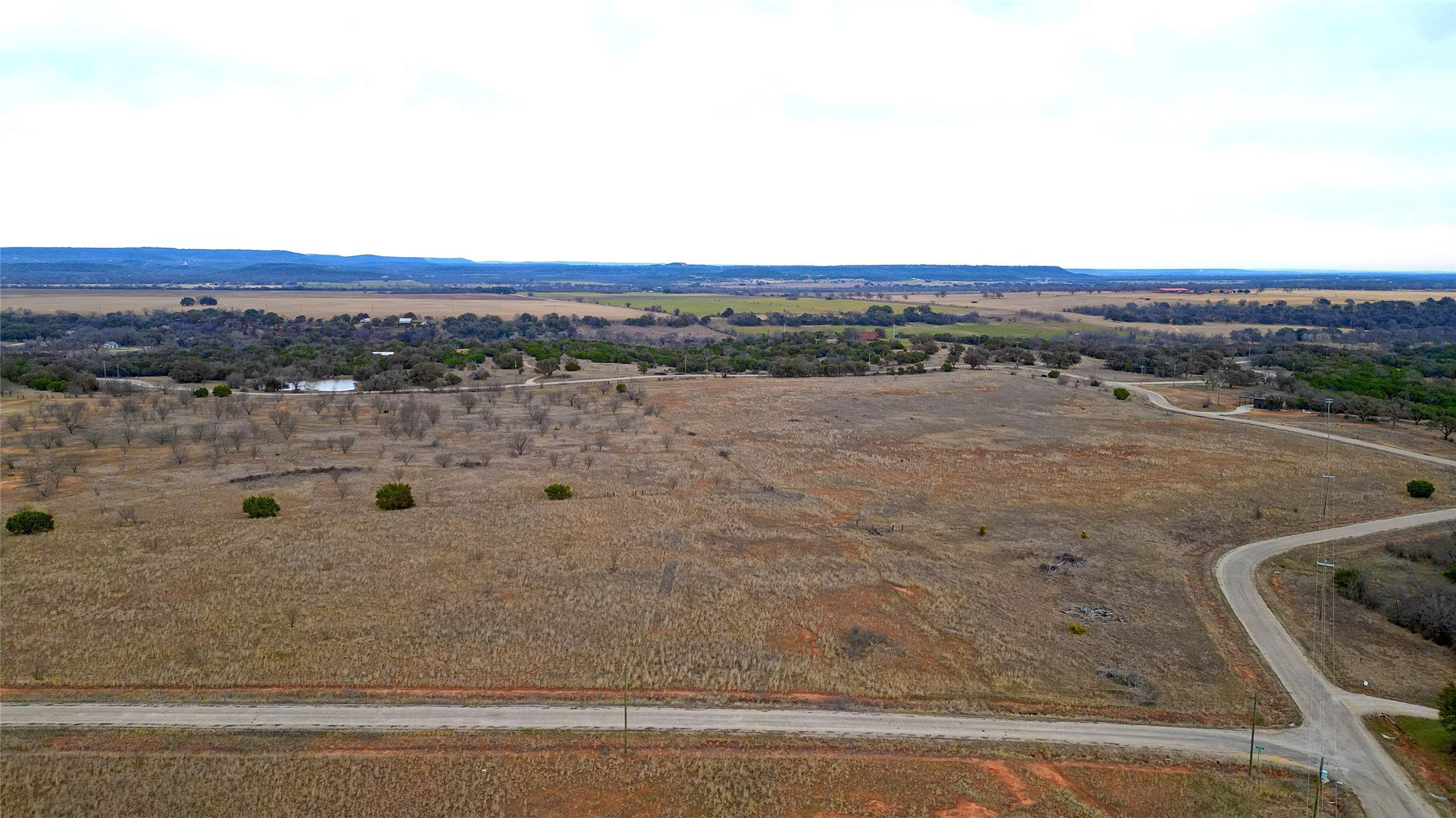 Aerial view of open rural land with scattered trees, a small pond, and roads.
