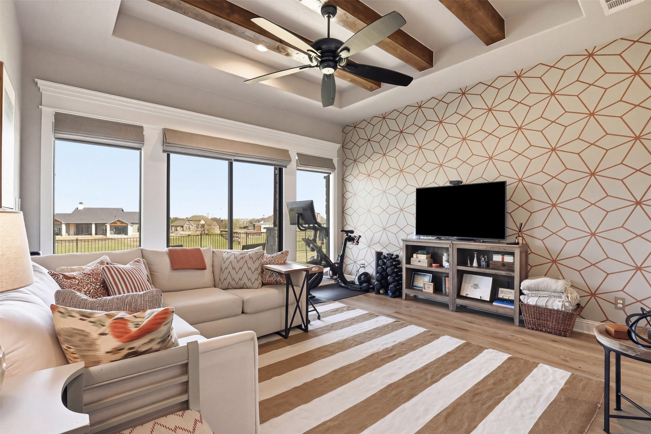 Living room with a beige sectional, geometric accent wall, workout equipment, and large windows.