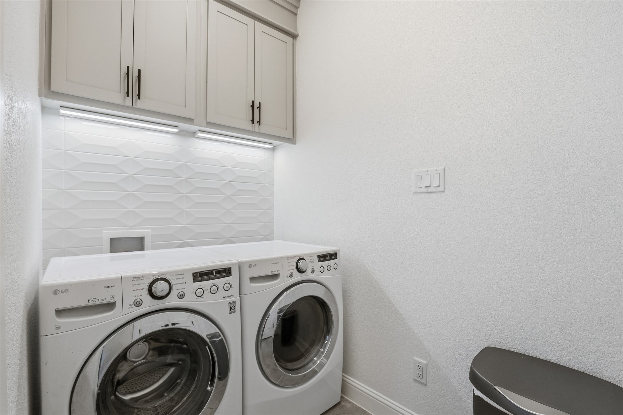 Laundry room with front-loading washer and dryer, white cabinets, and geometric tile backsplash.