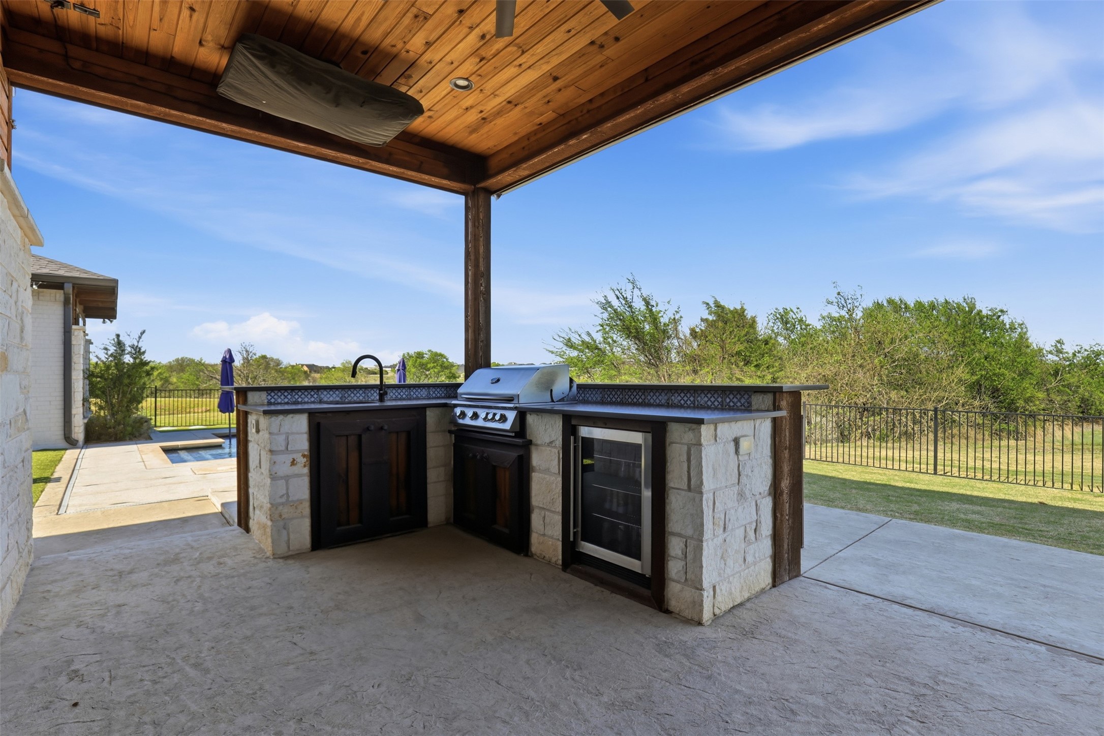 Covered stone outdoor kitchen with a built-in grill, sink, and beverage refrigerator beside the pool.