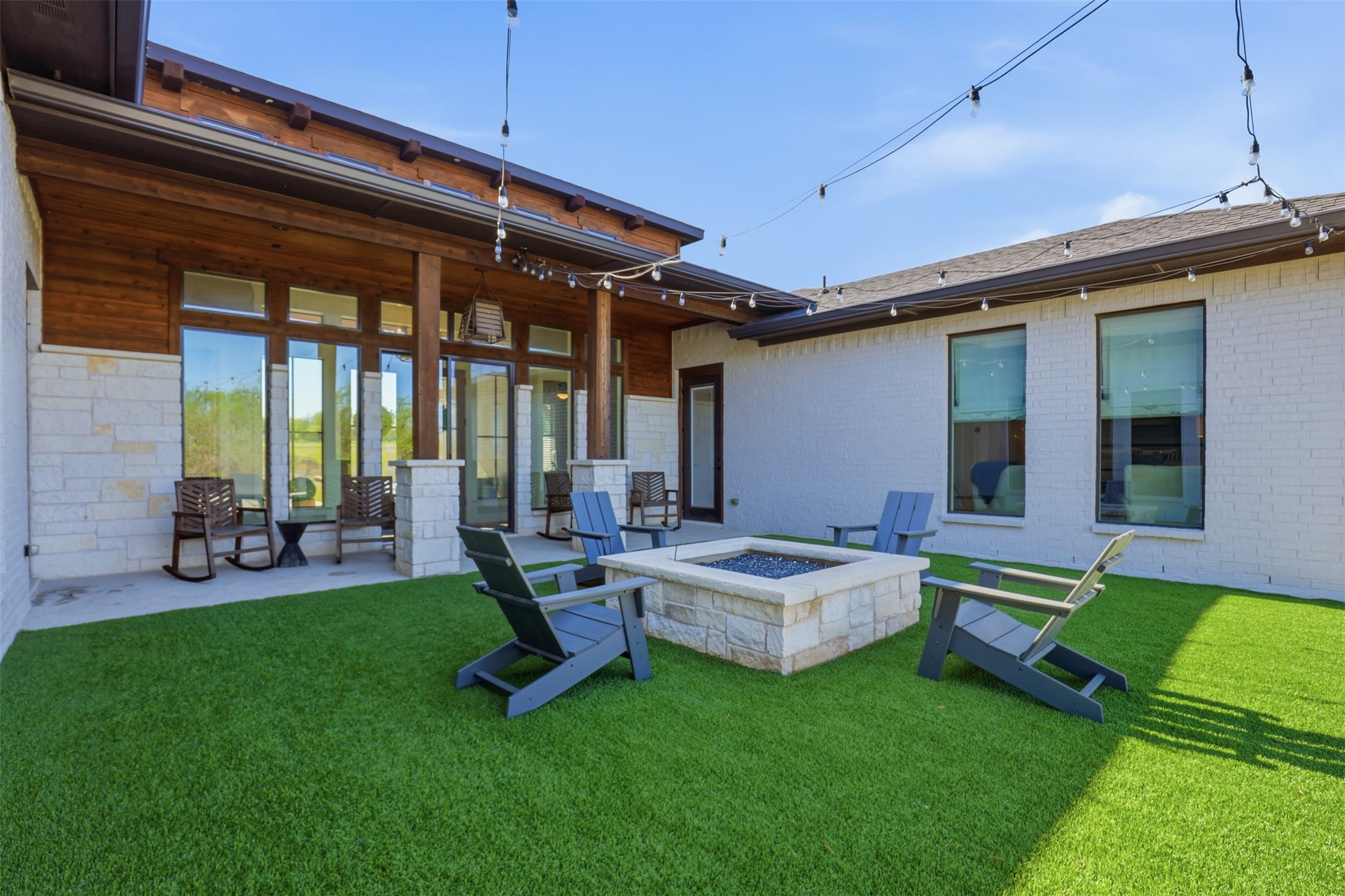 Backyard patio with a stone fire pit, Adirondack chairs, and overhead string lights.