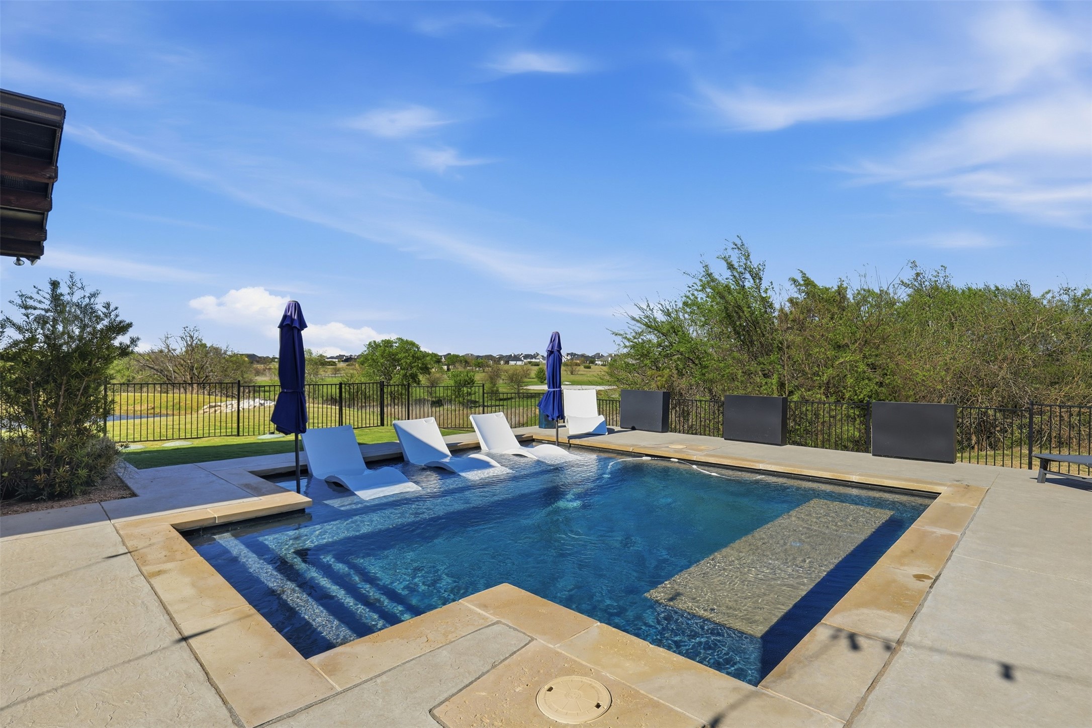 Backyard pool with a shallow tanning ledge, white loungers, and an open landscape view.