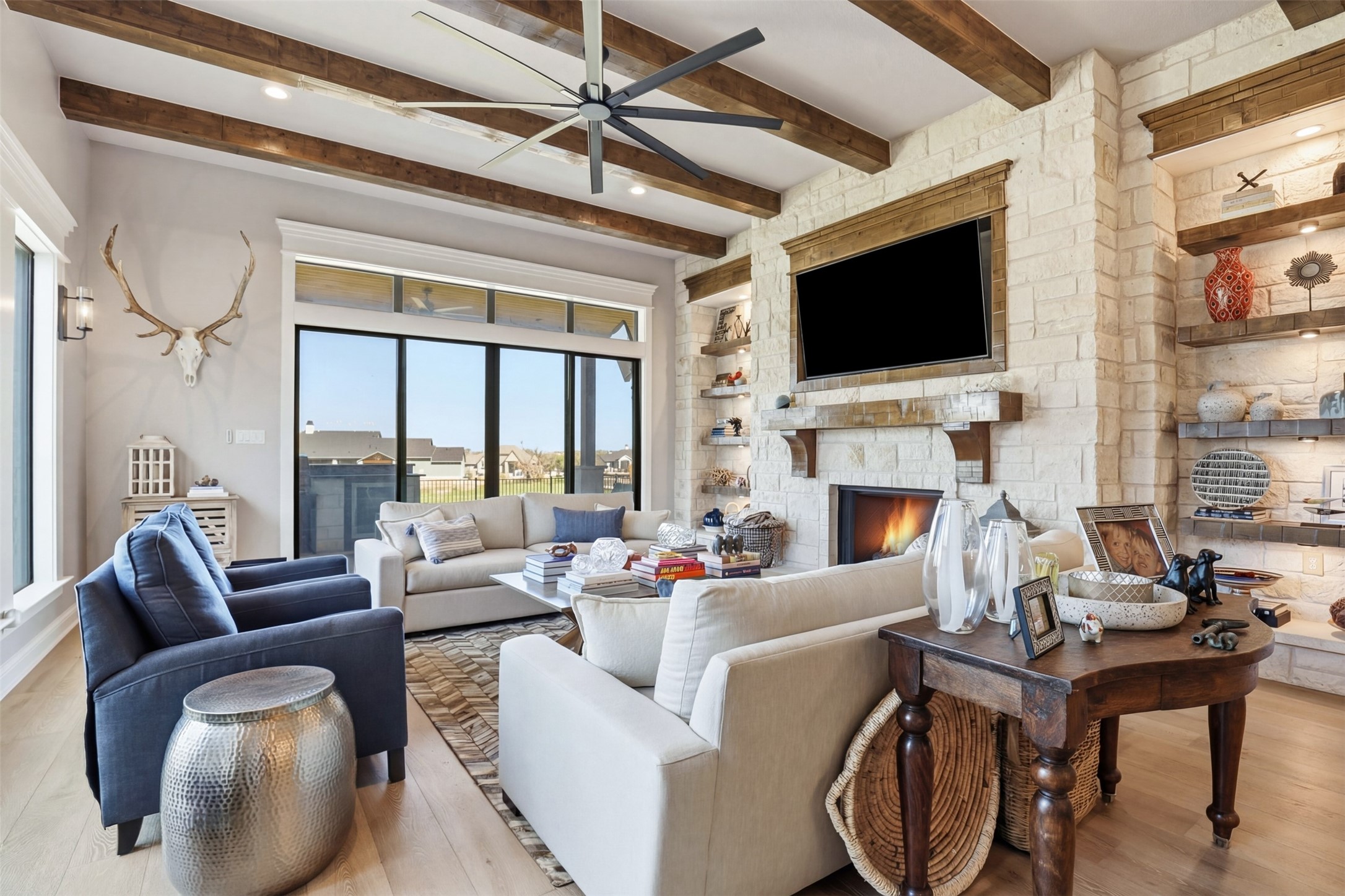 Living room with a tall stone fireplace, exposed ceiling beams, and large windows.